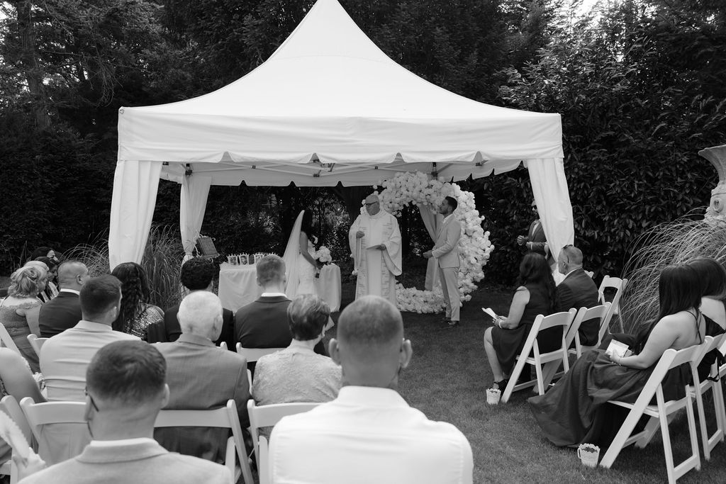 Outdoor wedding ceremony setup with white tent and chairs arranged on lawn under trees.