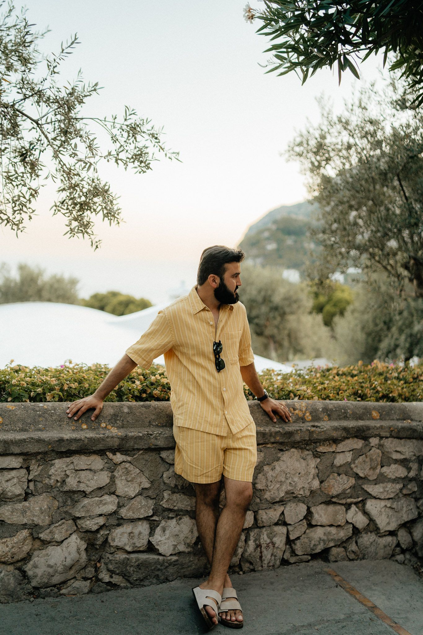 Person in yellow striped outfit leans against stone wall overlooking scenic Mediterranean landscape at sunset.