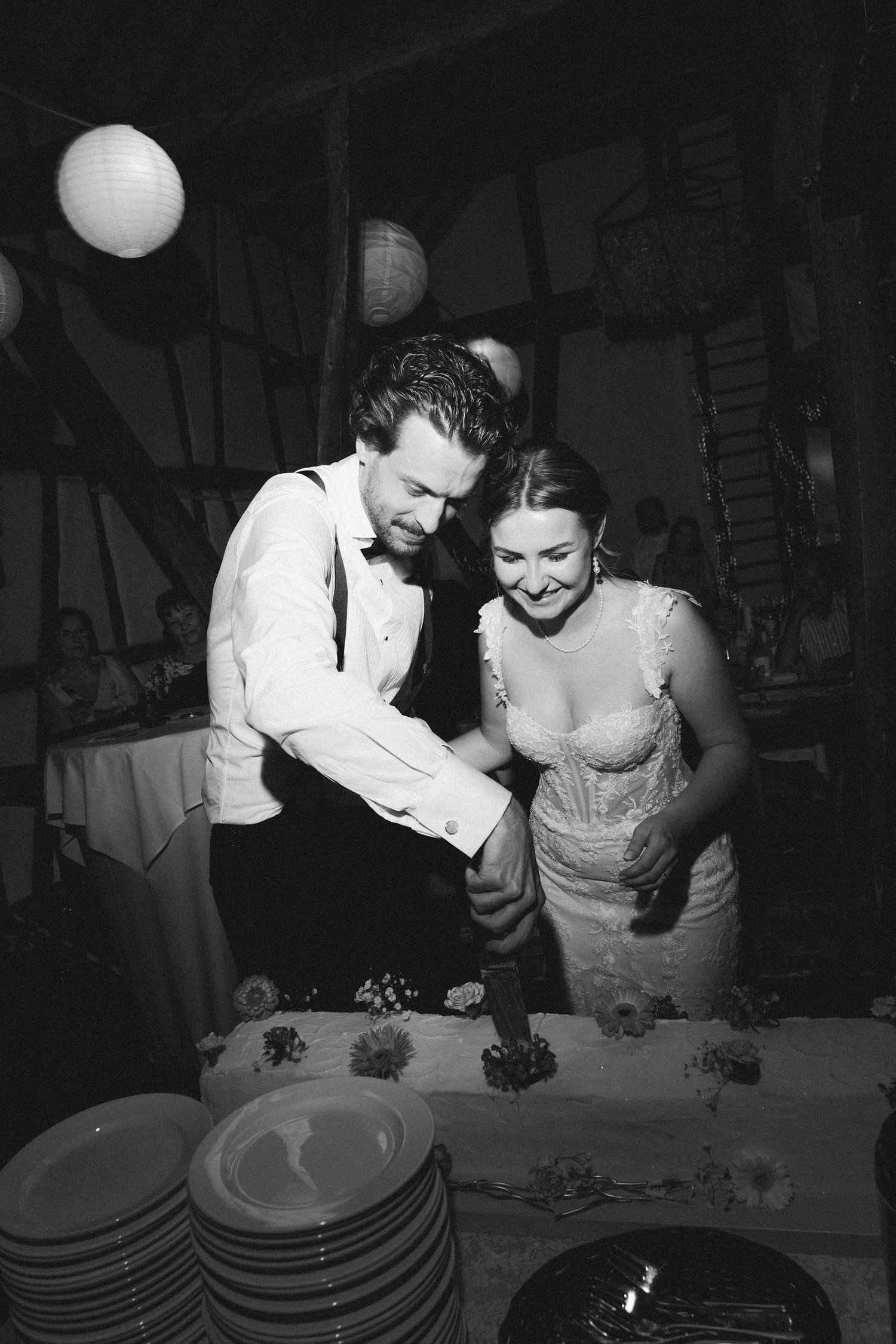 Black and white photo of bride and groom cutting wedding cake together, smiling, with paper lanterns in background.
