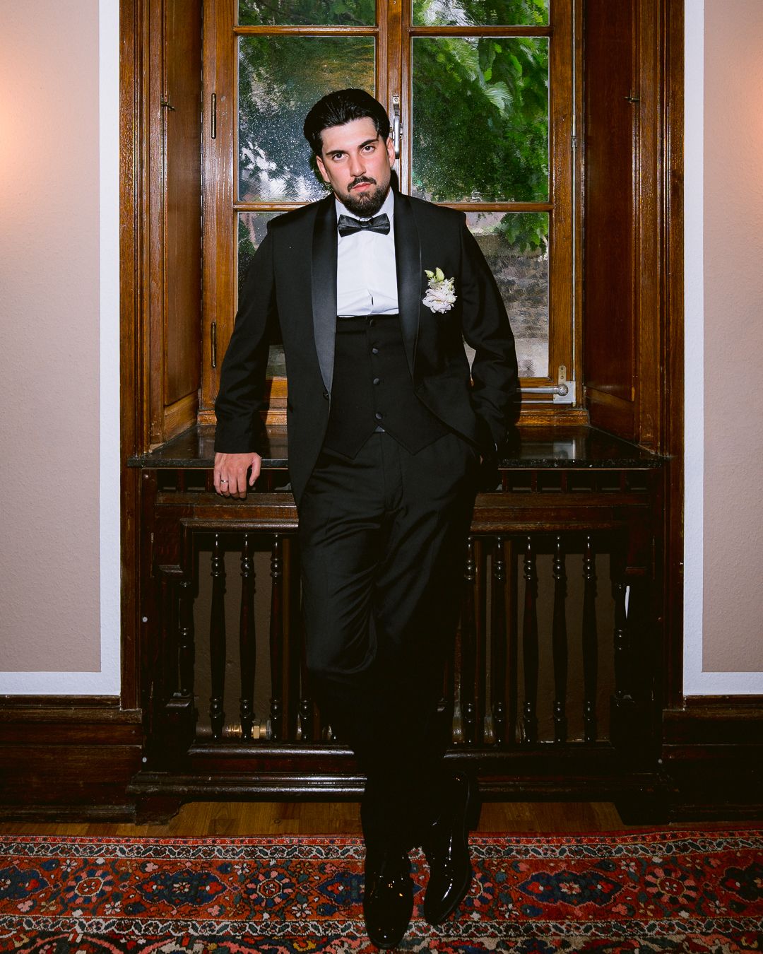 A groom in a black tuxedo with bow tie and boutonniere poses confidently in an elegant wood-paneled room.