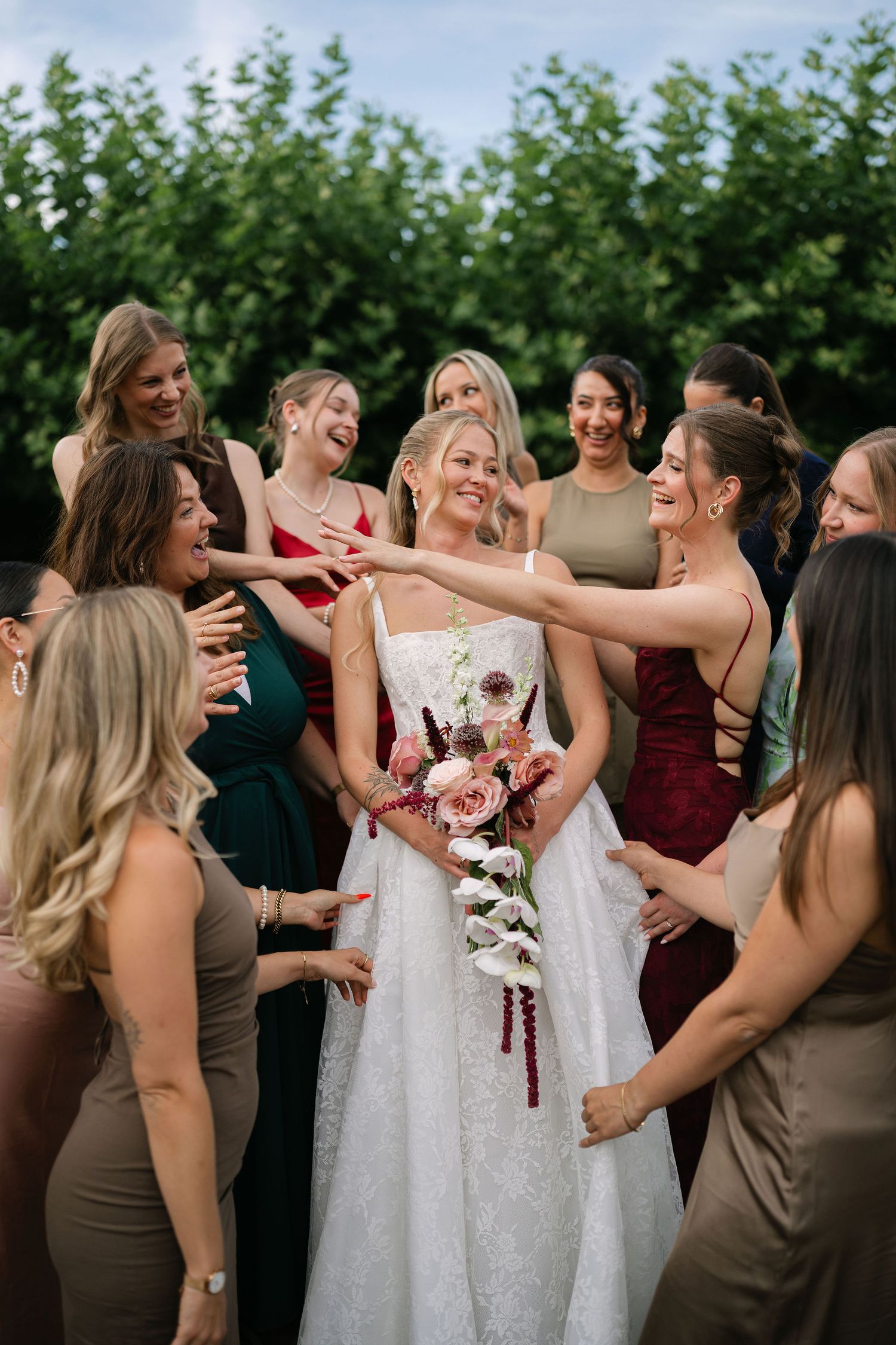 Bride in white lace gown surrounded by laughing bridesmaids in colorful dresses holding floral bouquet.