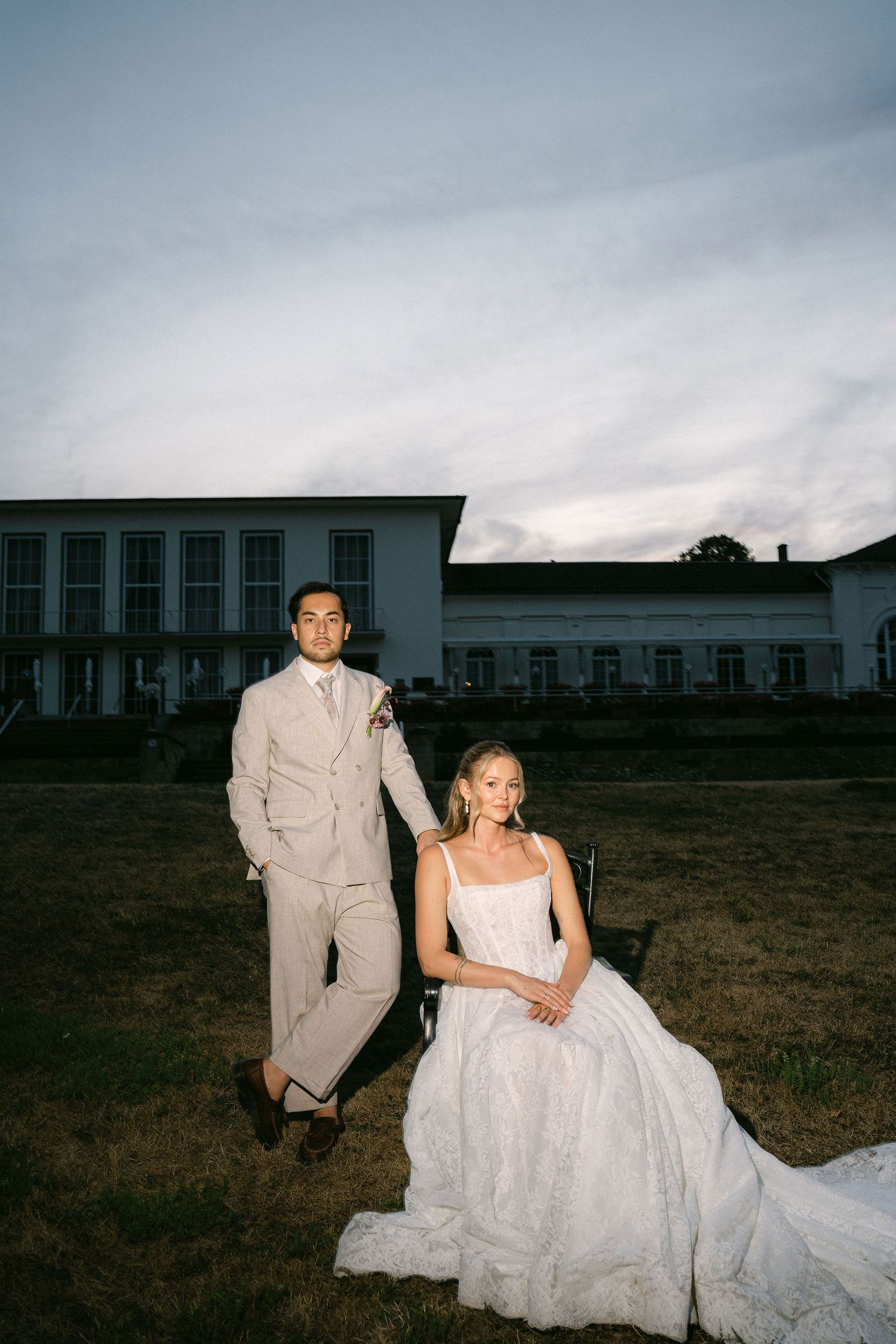 Bride in white gown seated outdoors with groom in beige suit standing beside her at dusk near elegant white venue.