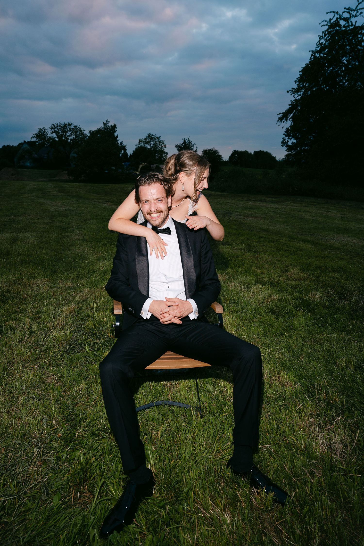 Joyful bride hugs groom from behind as he sits on chair in lush green field at dusk in formal attire.