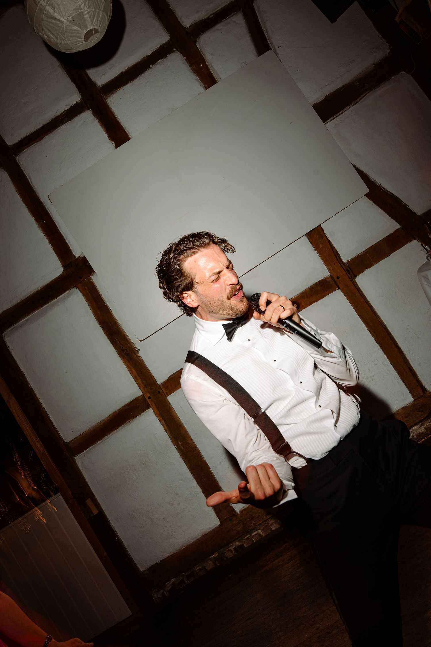 Man in white shirt, suspenders, and bow tie singing into a microphone at a wedding reception.