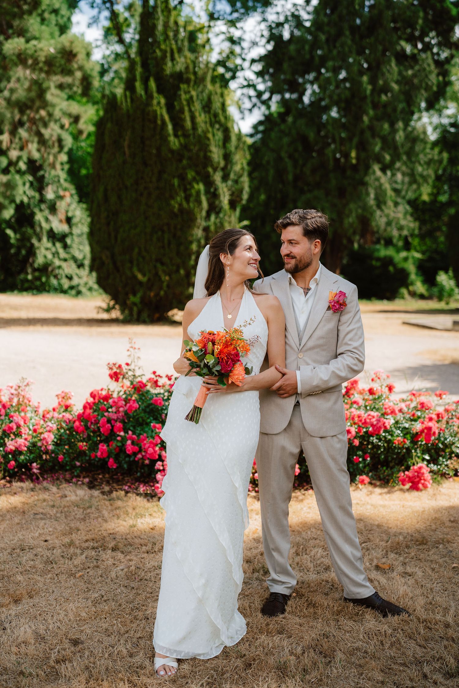 Bride and groom pose together in a garden with colorful flowers, she holds a vibrant bouquet in her white gown.