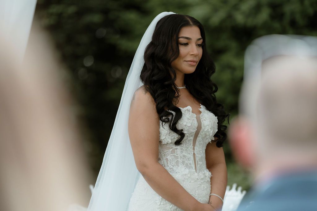 Beautiful bride in a white lace wedding dress with flowing veil smiles softly during outdoor ceremony.