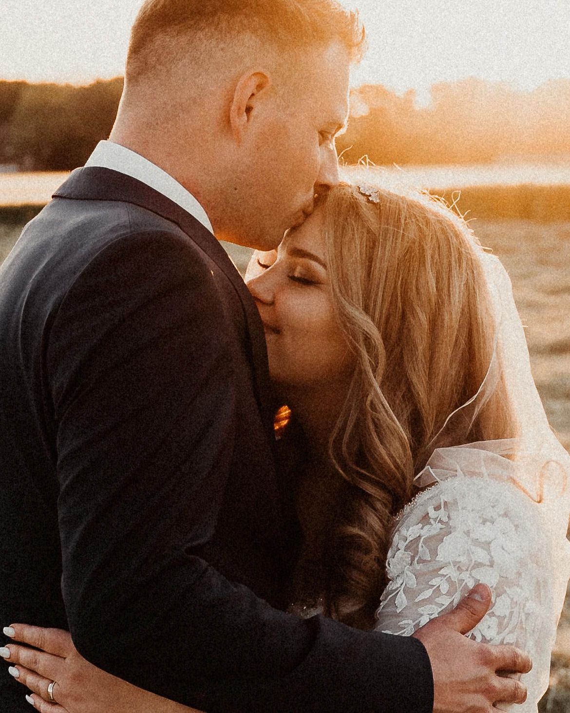 A romantic sunset wedding portrait of a couple embracing, with the bride wearing a lace dress and the groom in a dark suit.