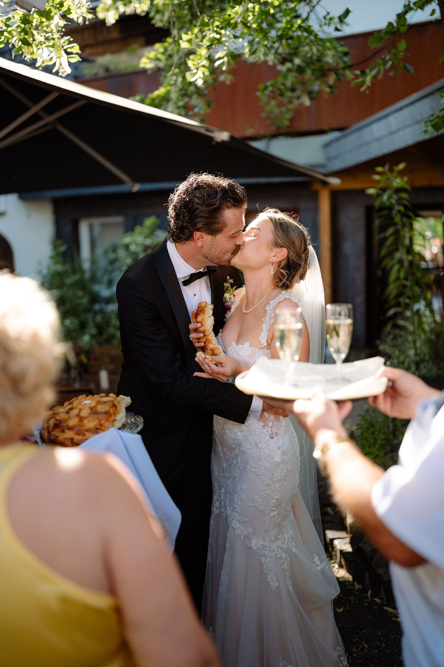 Bride and groom share a kiss outdoors at their wedding reception, holding champagne and food in sunny garden setting.