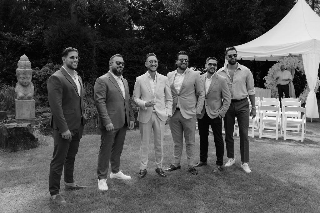 A line of formally dressed wedding guests stand outdoors next to white folding chairs at an outdoor ceremony venue.