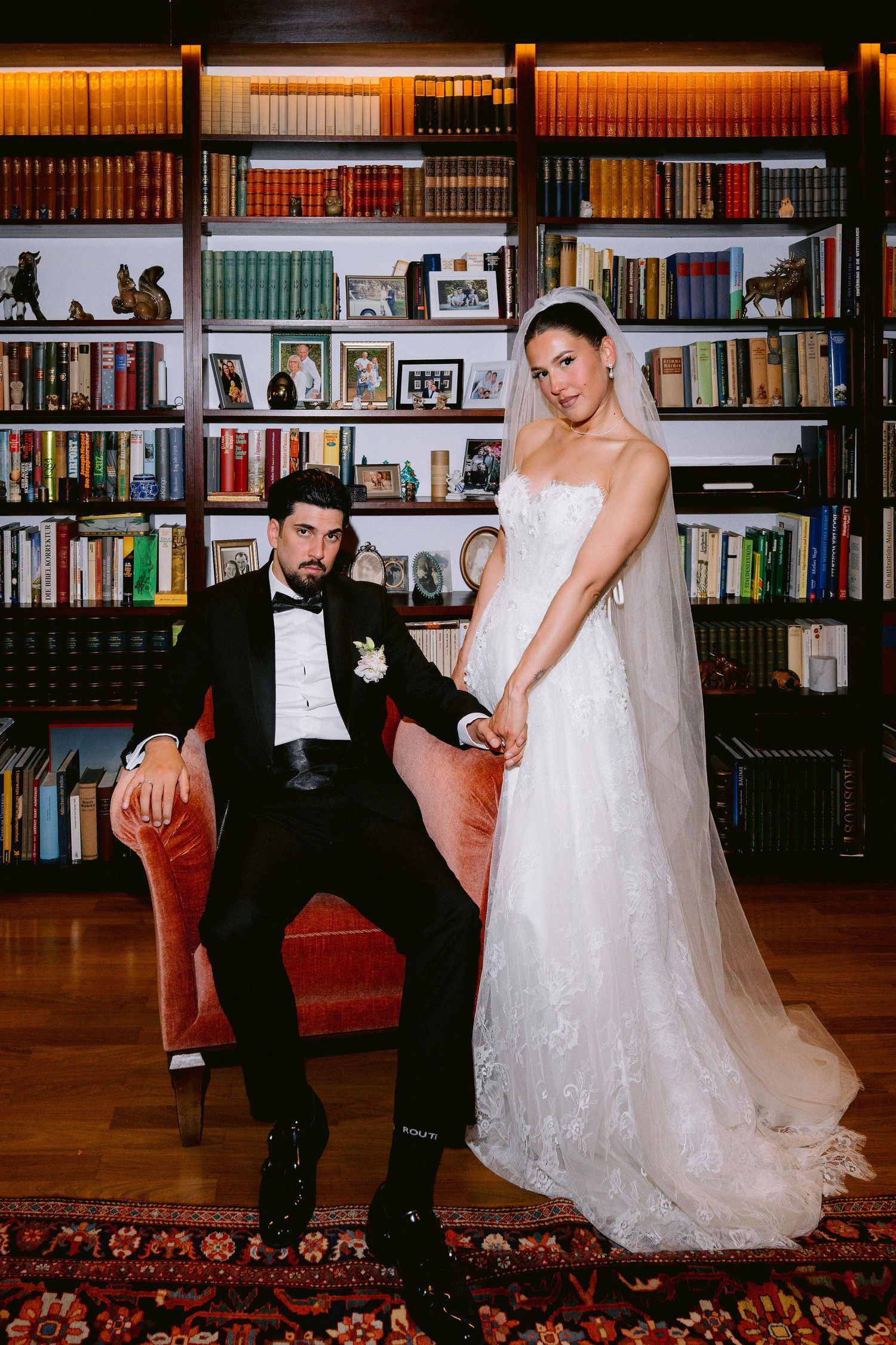 Elegant bride and groom pose in a library, she in lace gown with veil, he in black tuxedo seated in velvet chair.