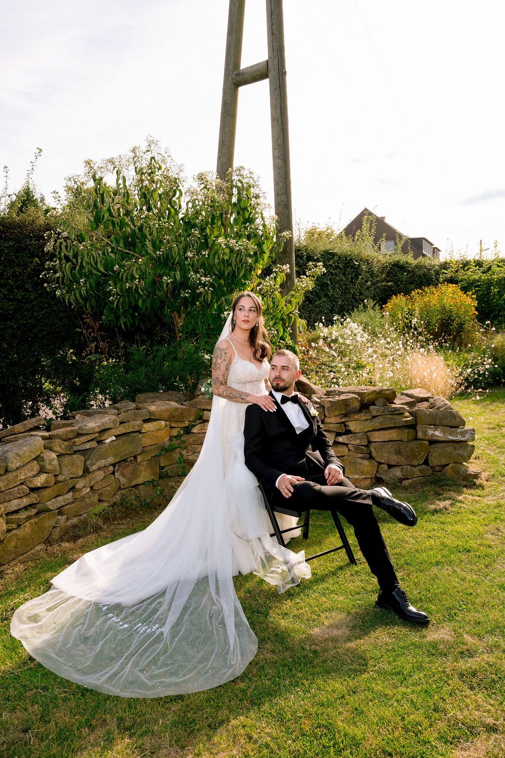 A couple in wedding attire share romantic moments on a grassy hillside with a stone wall backdrop during their outdoor photoshoot.