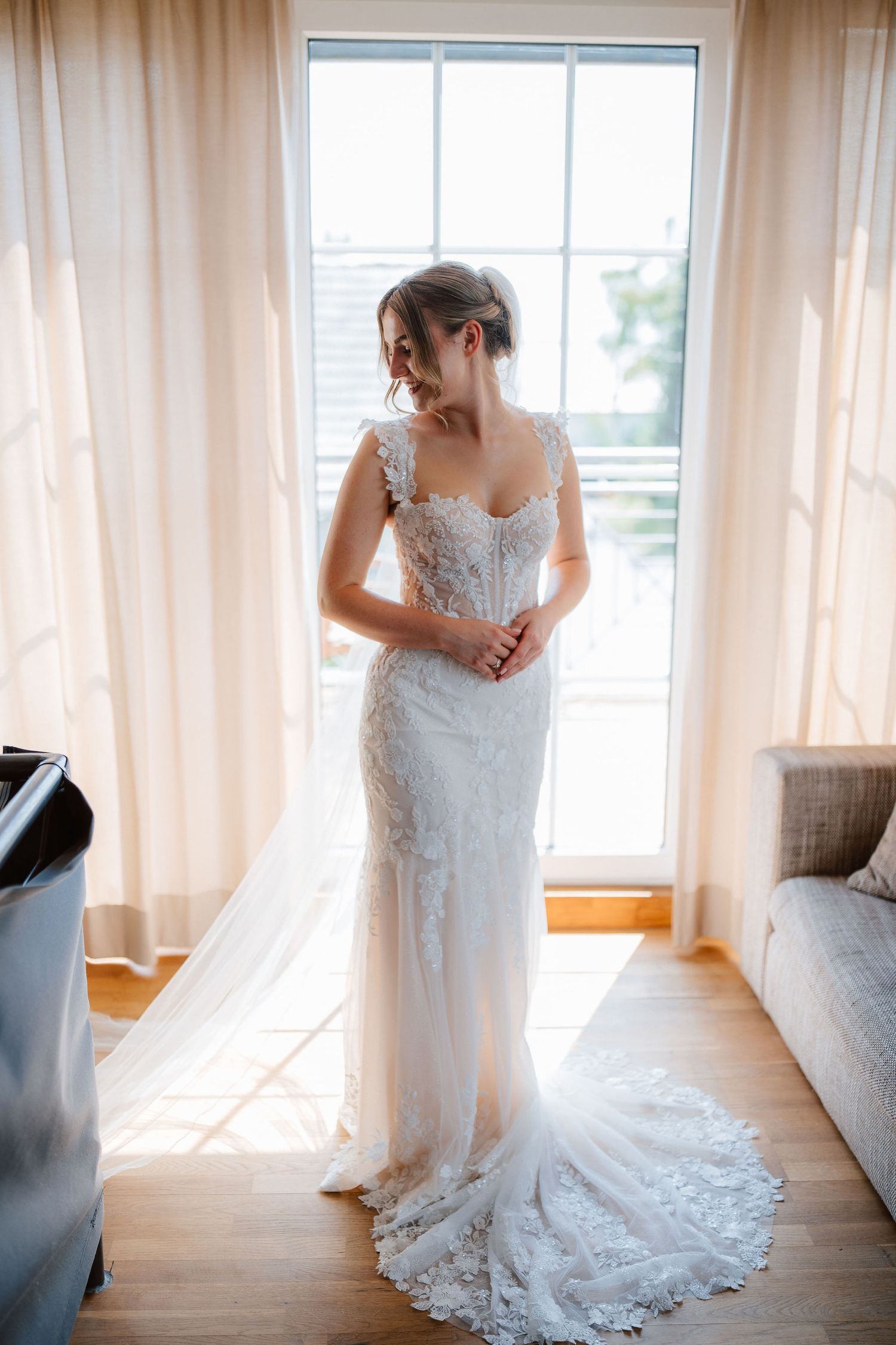 Bride in elegant lace wedding gown standing by sunlit window, showcasing intricate details and long train.