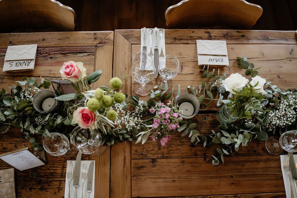 Rustic wooden table decorated with elegant pink roses, flowing eucalyptus garland and crystal wine glasses for dining.