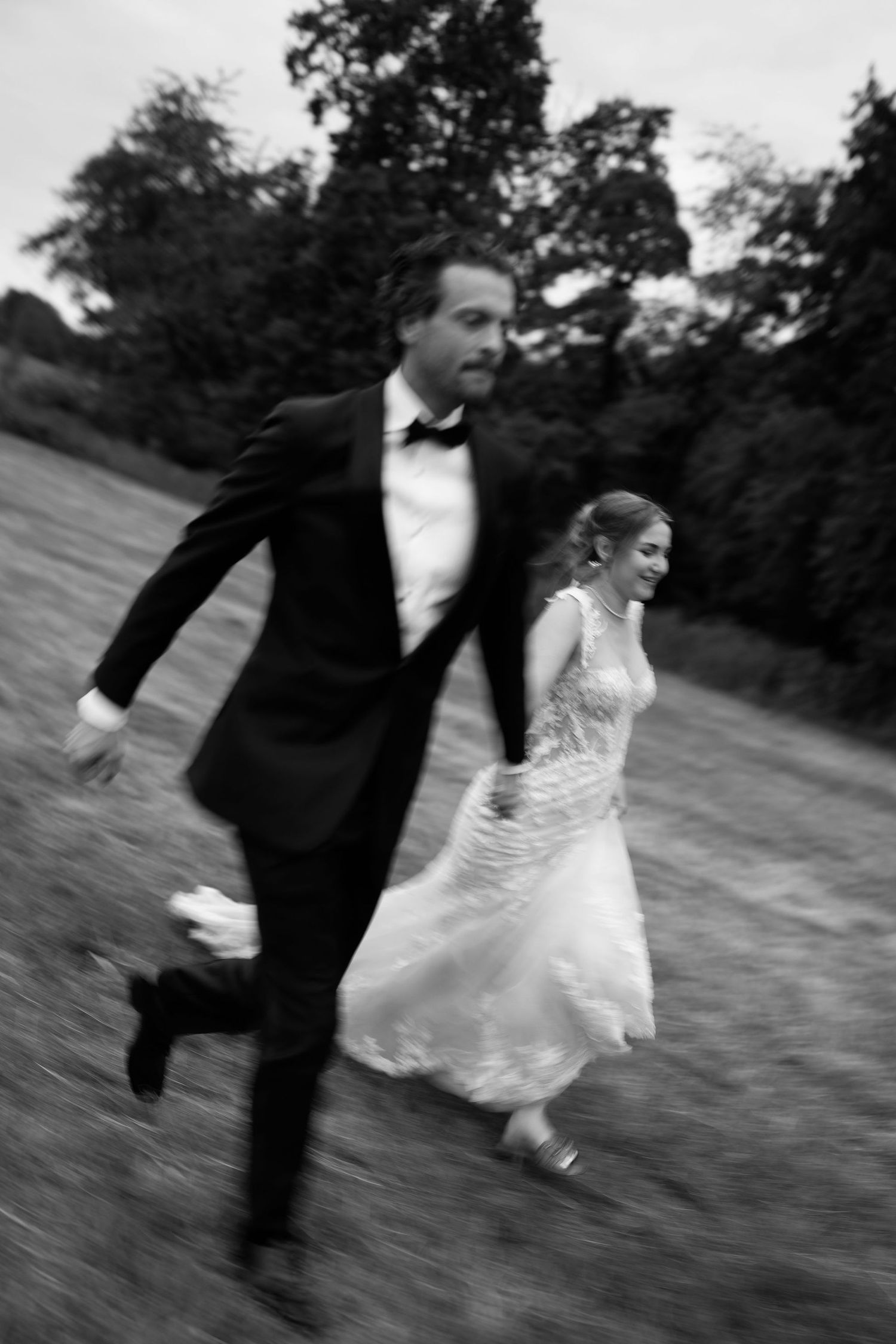 Black and white photo of a bride and groom joyfully running through a field, holding hands in wedding attire.
