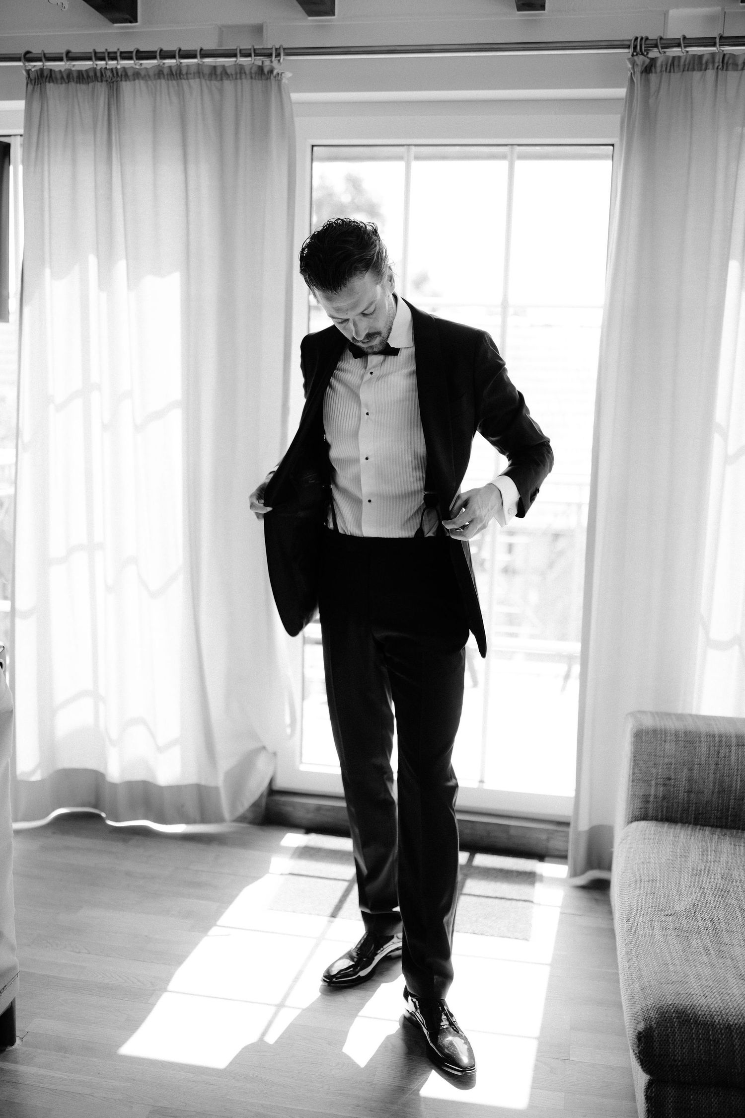Groom in black tuxedo adjusts jacket while standing in sunlit room in black and white photo.
