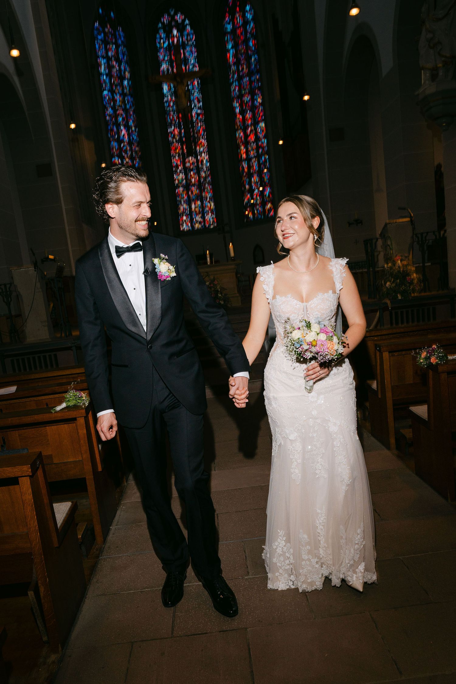 Bride and groom walk hand-in-hand down church aisle, bride in lace gown holding bouquet, groom in black tuxedo.