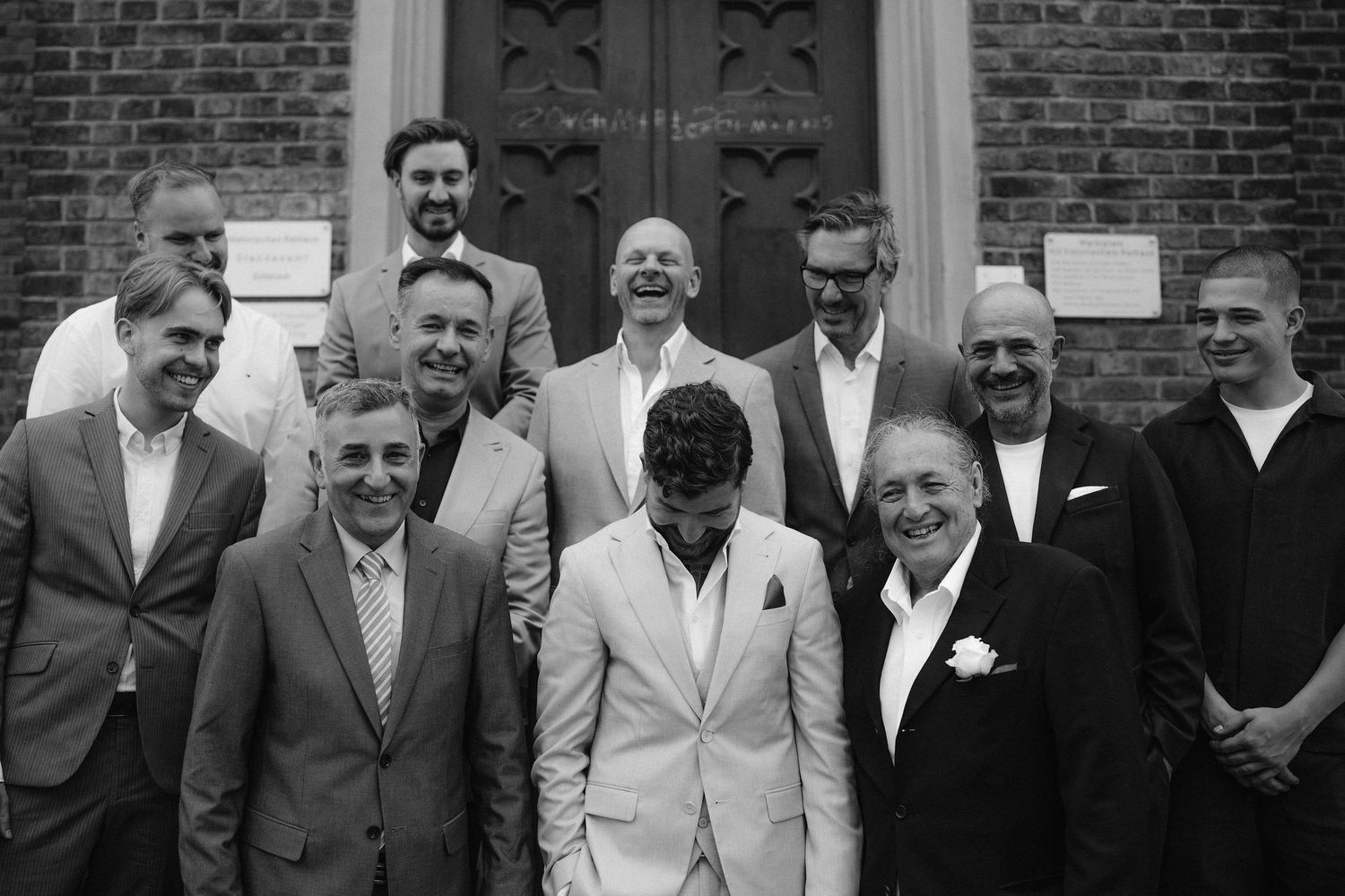 Black and white photo of a group of men in suits posing together outside a brick building for a wedding.
