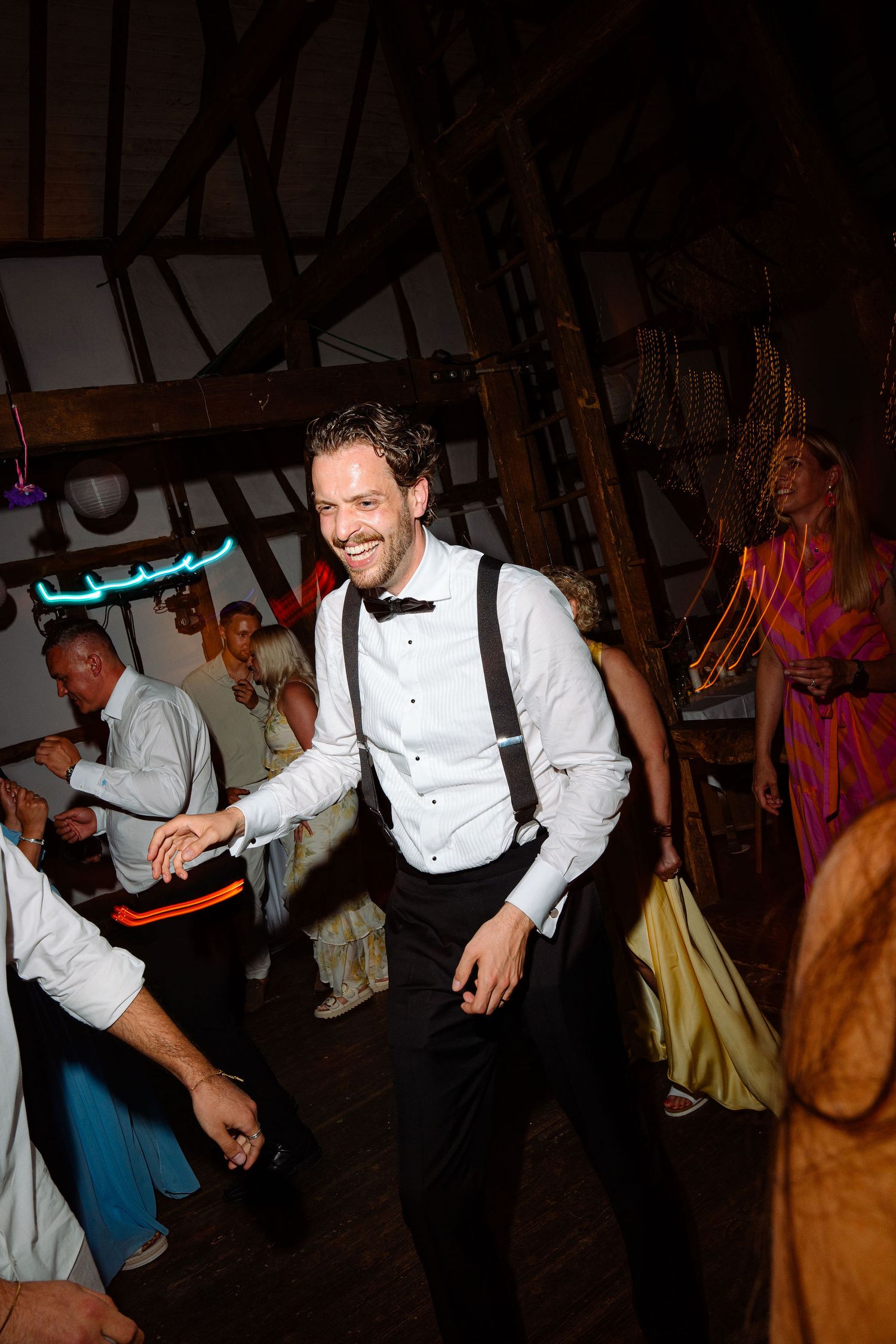 A smiling groom in suspenders and bow tie dances joyfully at a rustic barn wedding reception.
