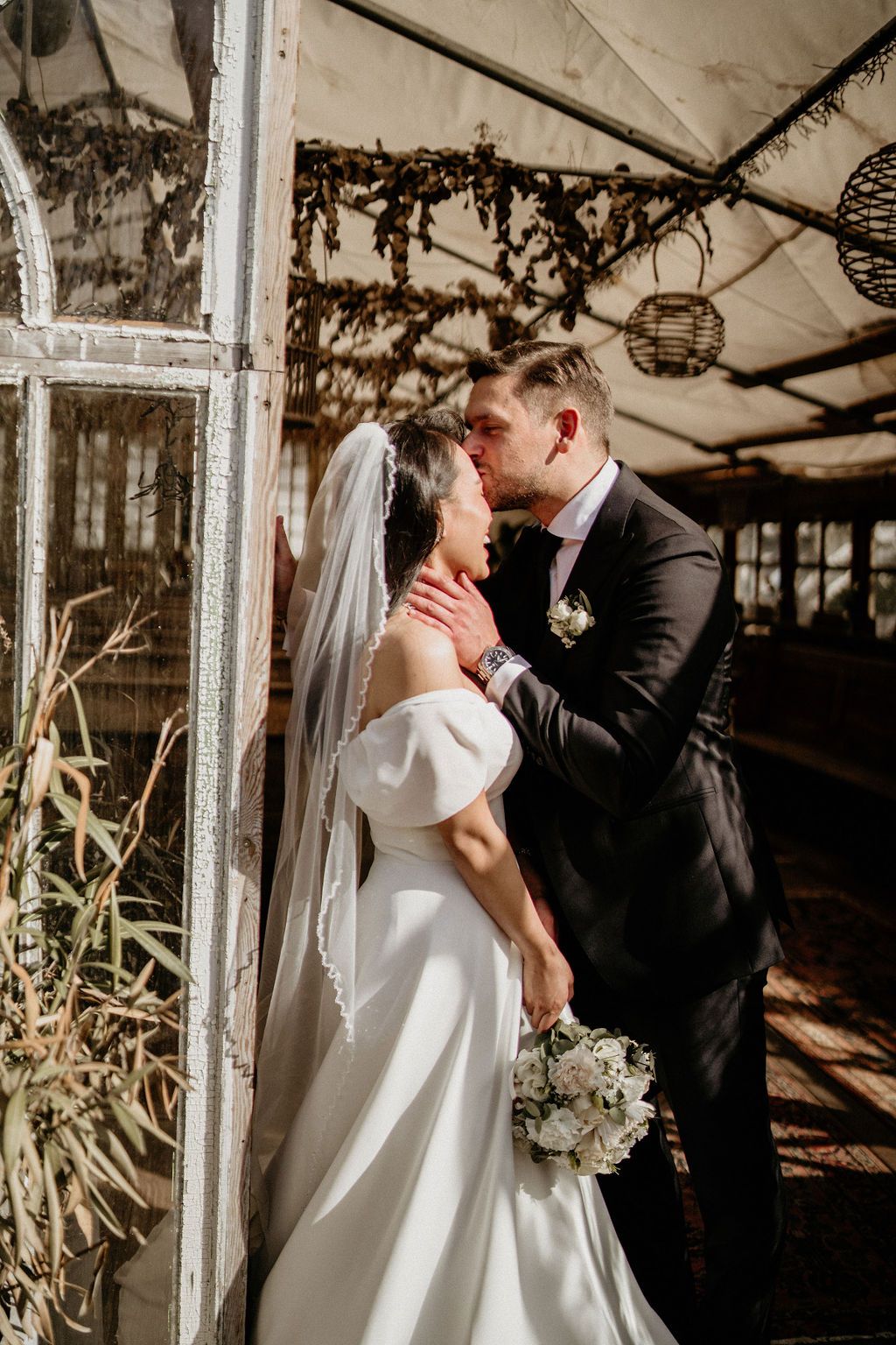 A romantic wedding moment captured in a rustic greenhouse with hanging lanterns and sunlit ambiance.