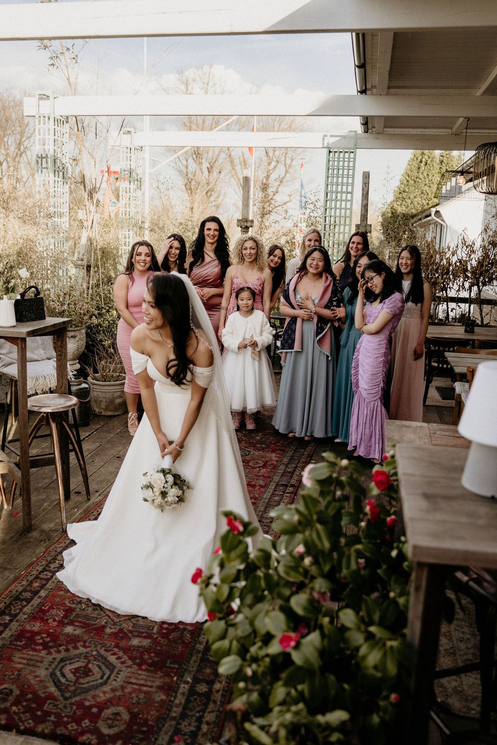 A joyful wedding party in mix-and-match pastel bridesmaid dresses gather for photos in a rustic indoor venue.