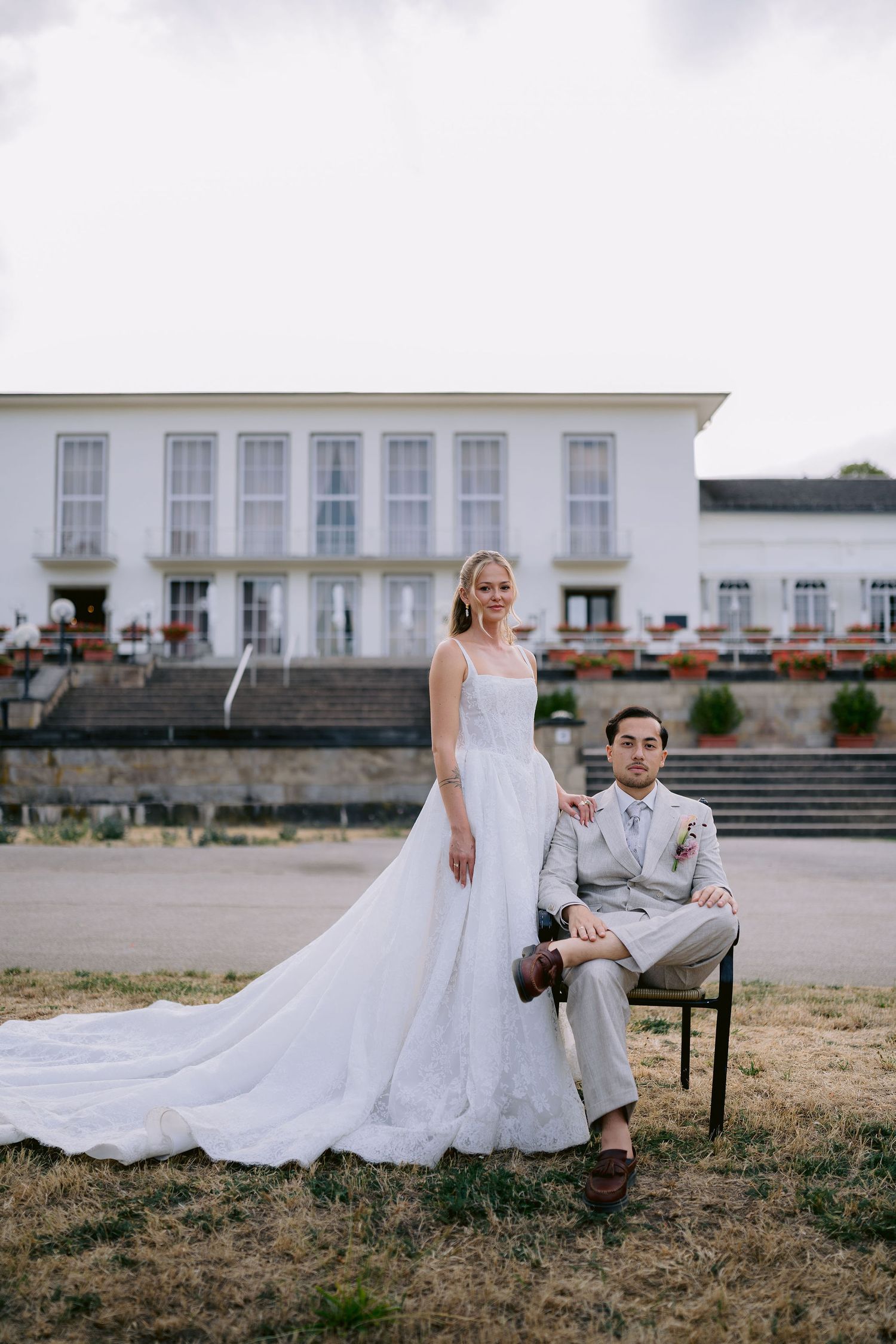 Bride in white gown stands beside groom in gray suit seated on chair outside elegant white venue.
