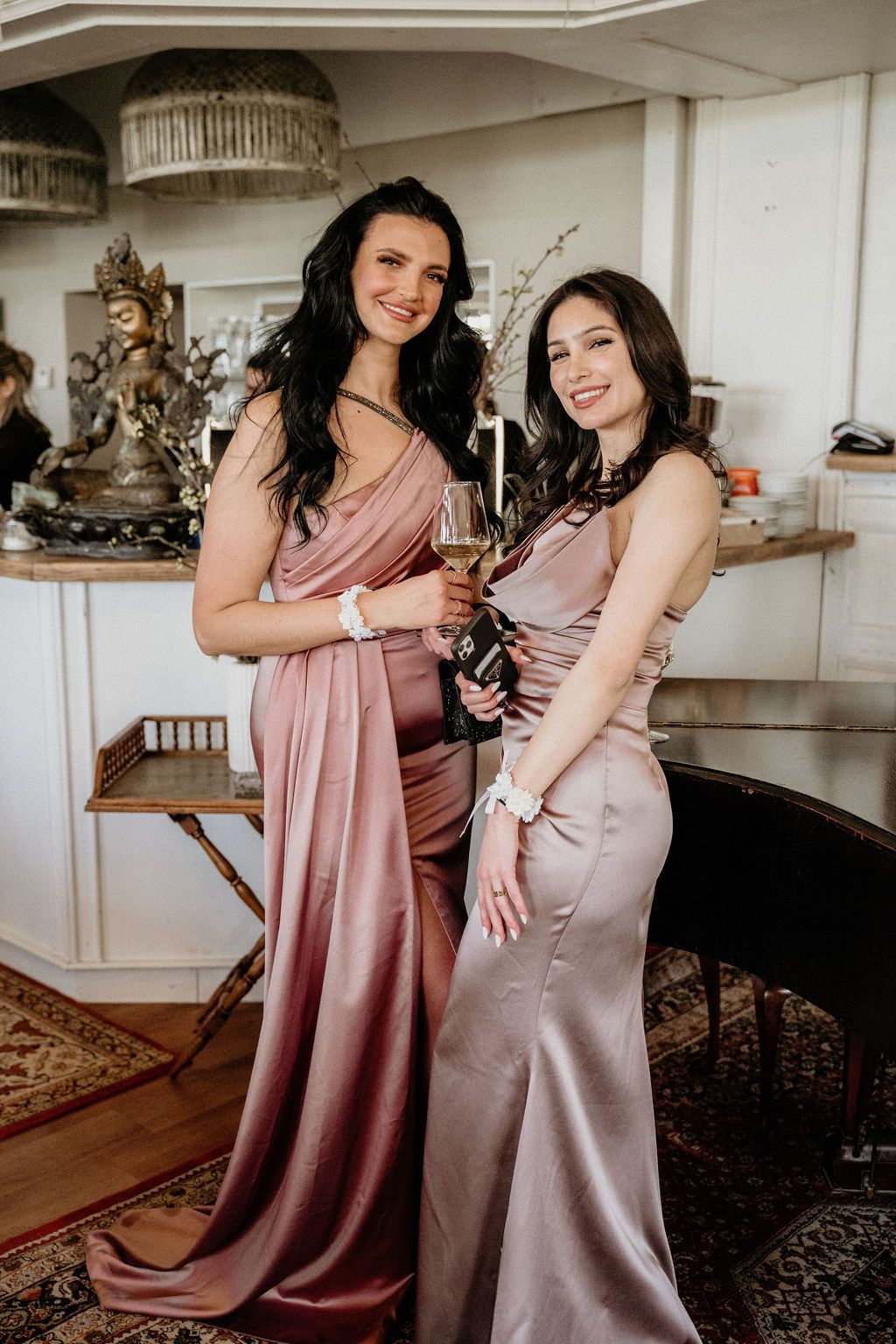 Two bridesmaids pose together in elegant silk gowns in mauve and dusty rose colors at an indoor event.