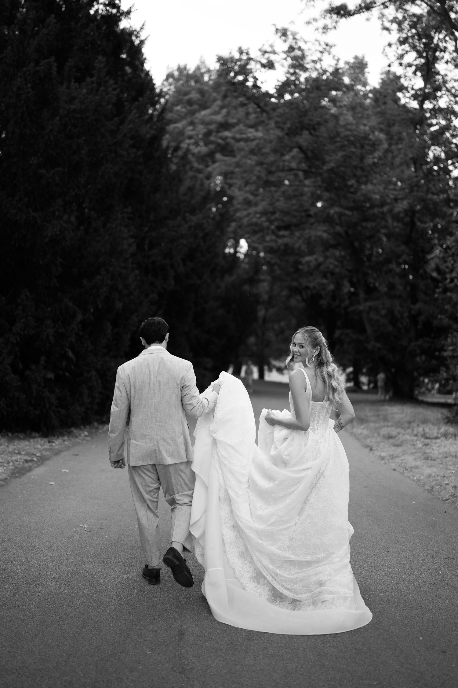 Bride and groom walking hand in hand down tree-lined path in romantic black and white wedding photo.