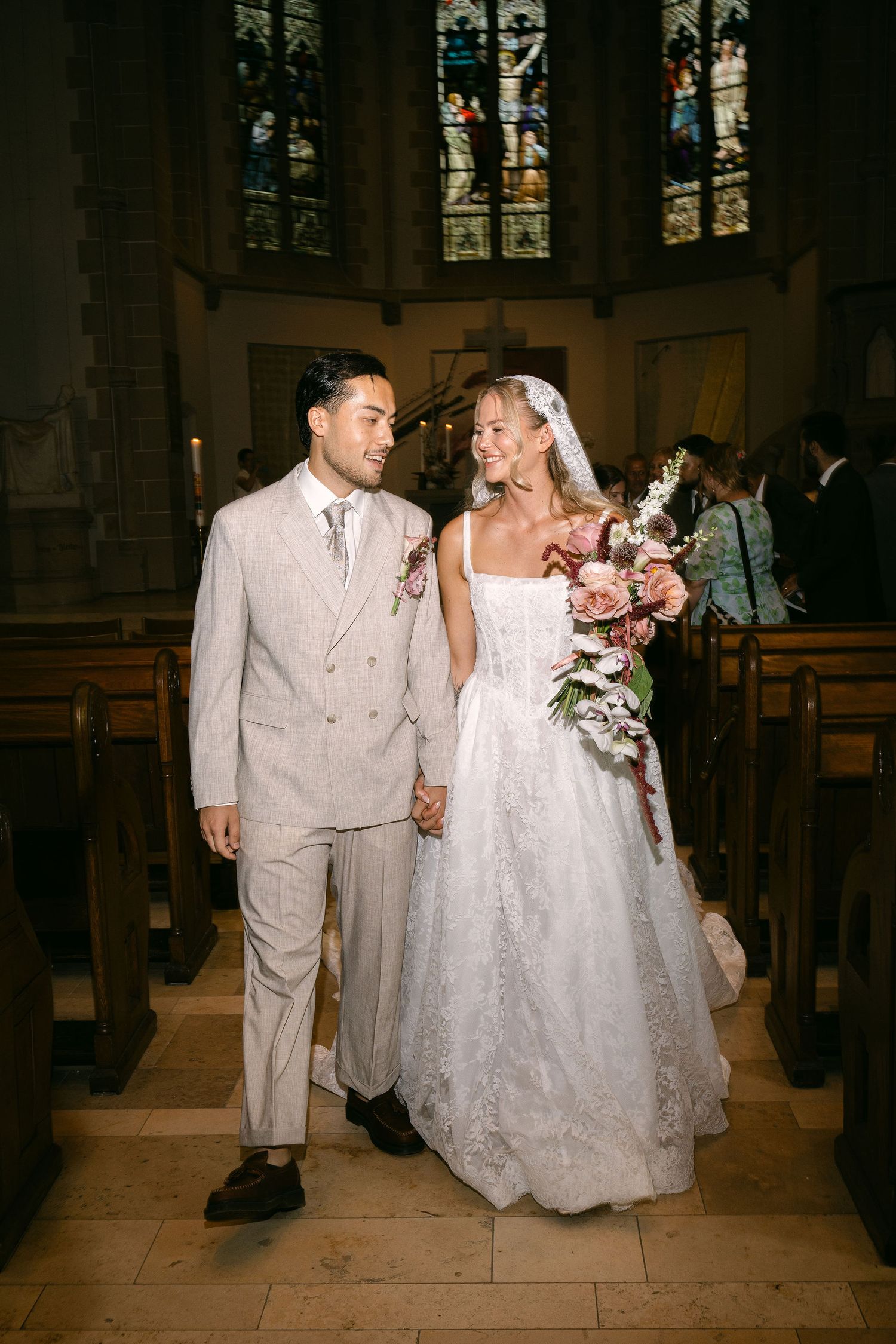 Bride and groom walk down church aisle, she in white lace gown with veil, he in beige suit, holding hands.