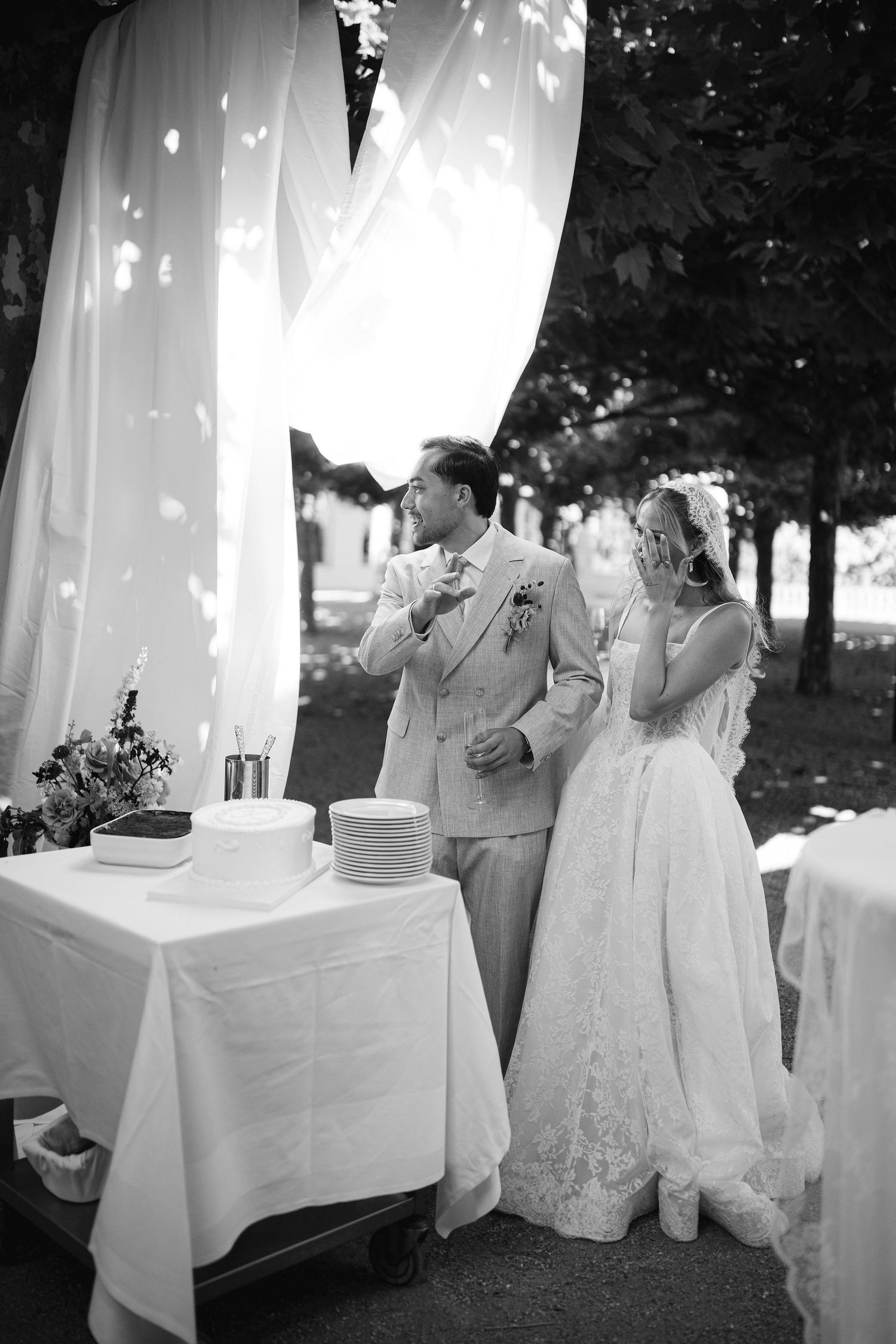 Bride and groom laugh together while cutting their wedding cake at an outdoor reception.