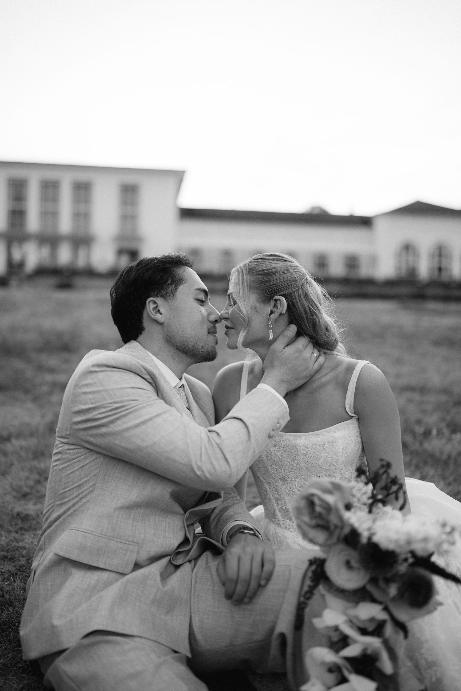 Black and white photo of a groom tenderly touching his bride's face while sitting on grass outside a venue.