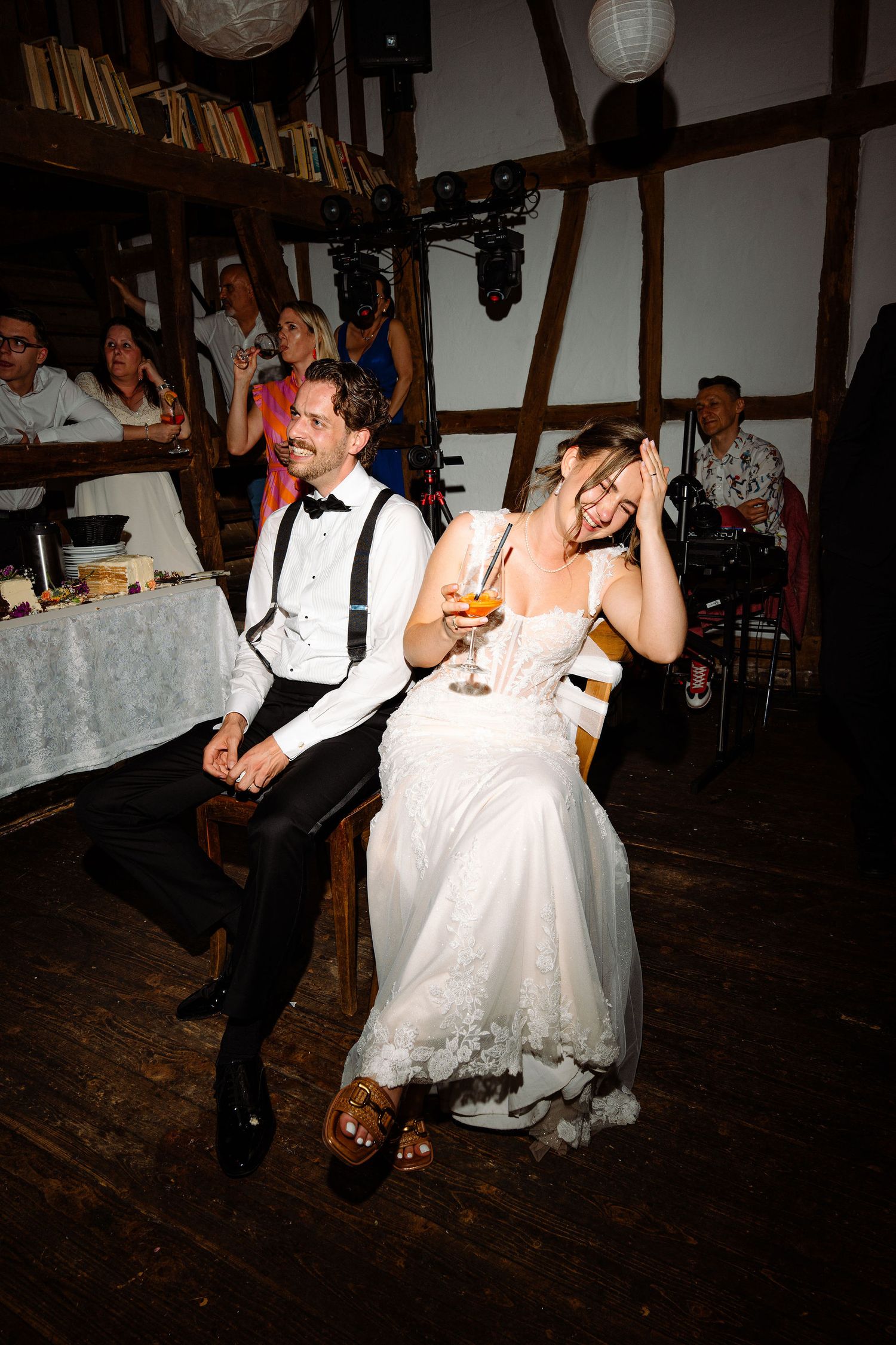 Bride and groom laugh joyfully at wedding reception in rustic barn venue with guests in background.