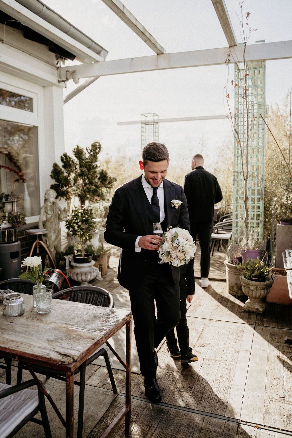 Black-suited figure holding a floral bouquet in a rustic sunlit venue with wooden floors and potted plants.