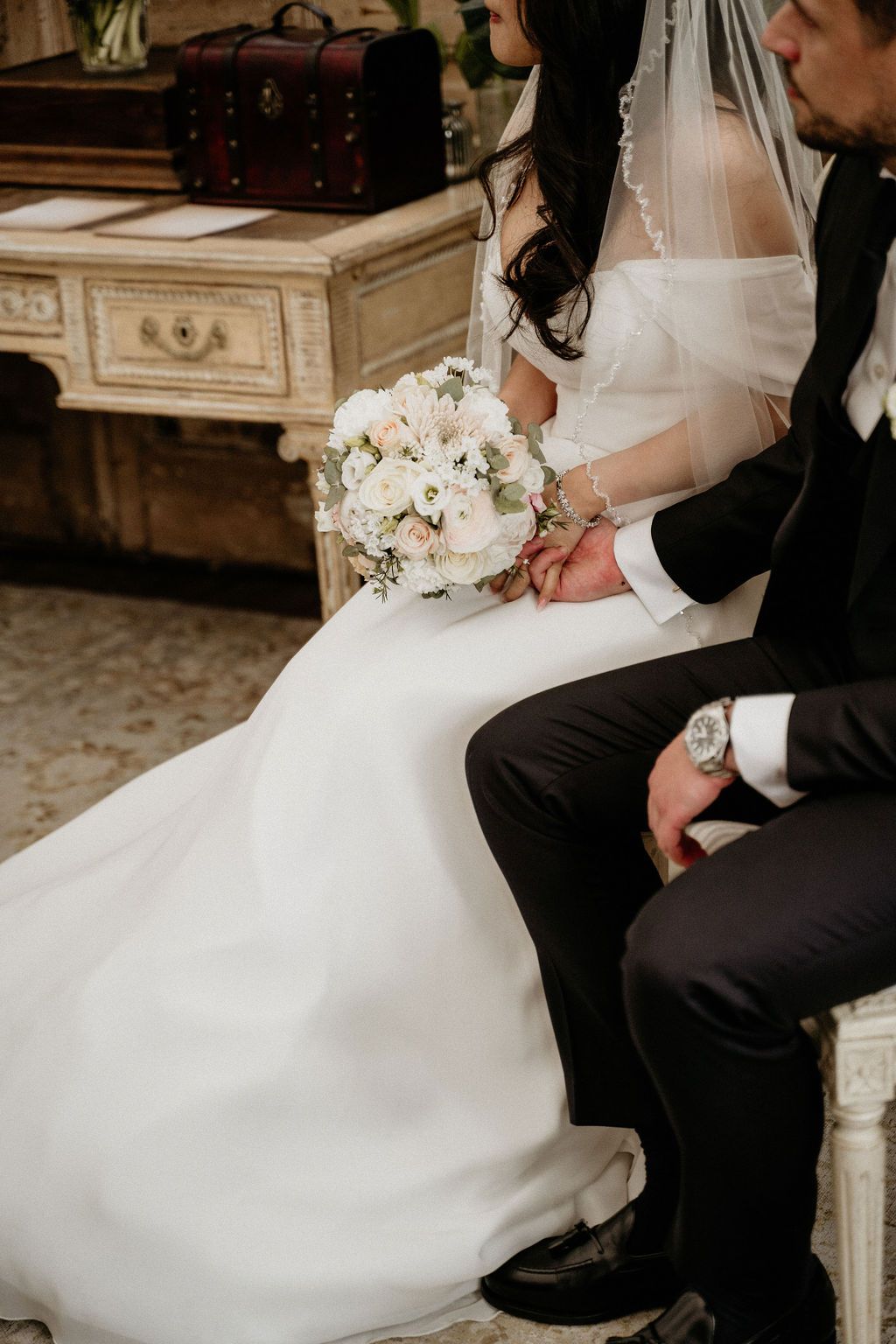 A wedding couple sits together holding a white floral bouquet with ornate vintage furniture in the background.