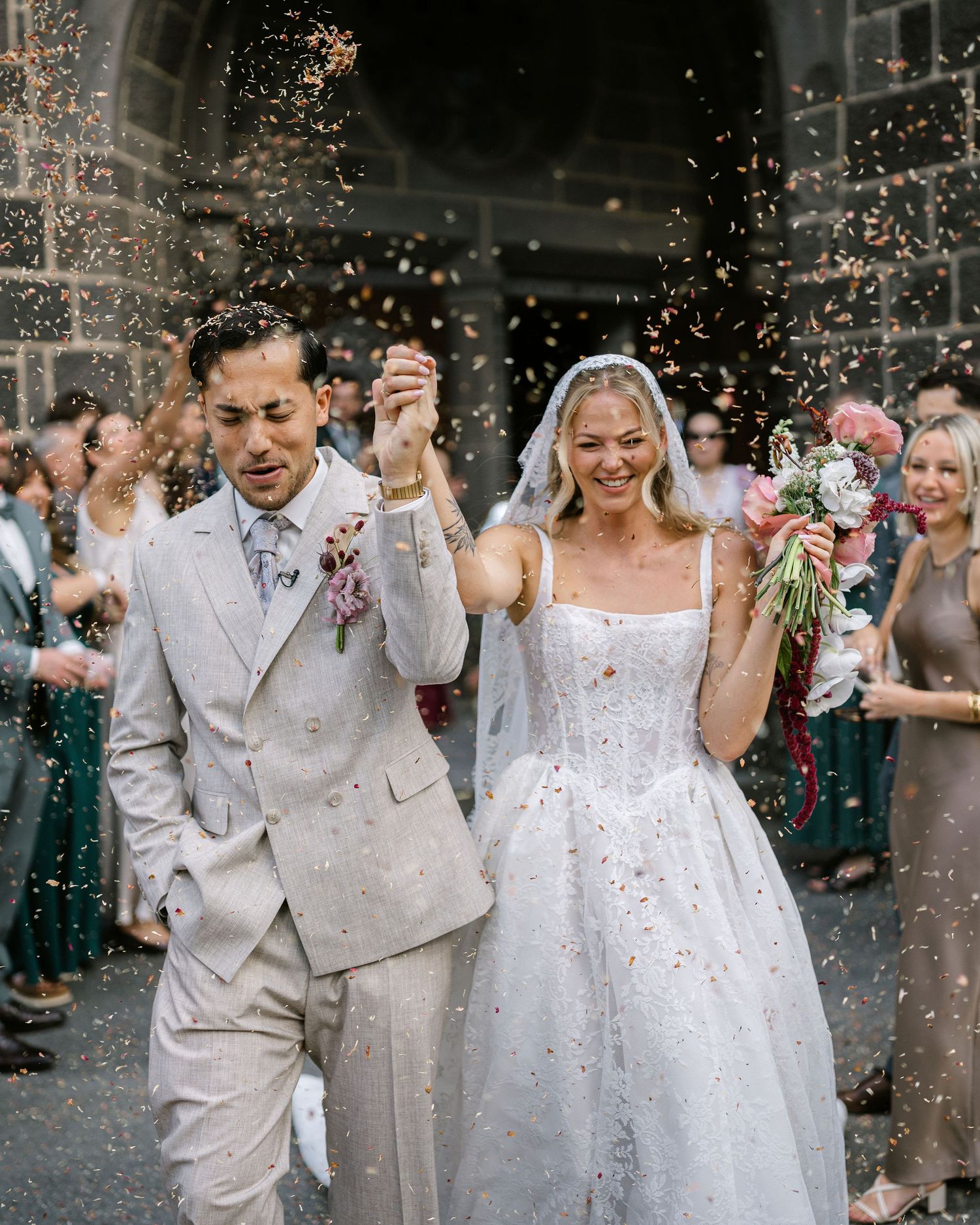 Joyful newlyweds exit church in confetti shower, bride in white gown with veil, groom in beige suit.