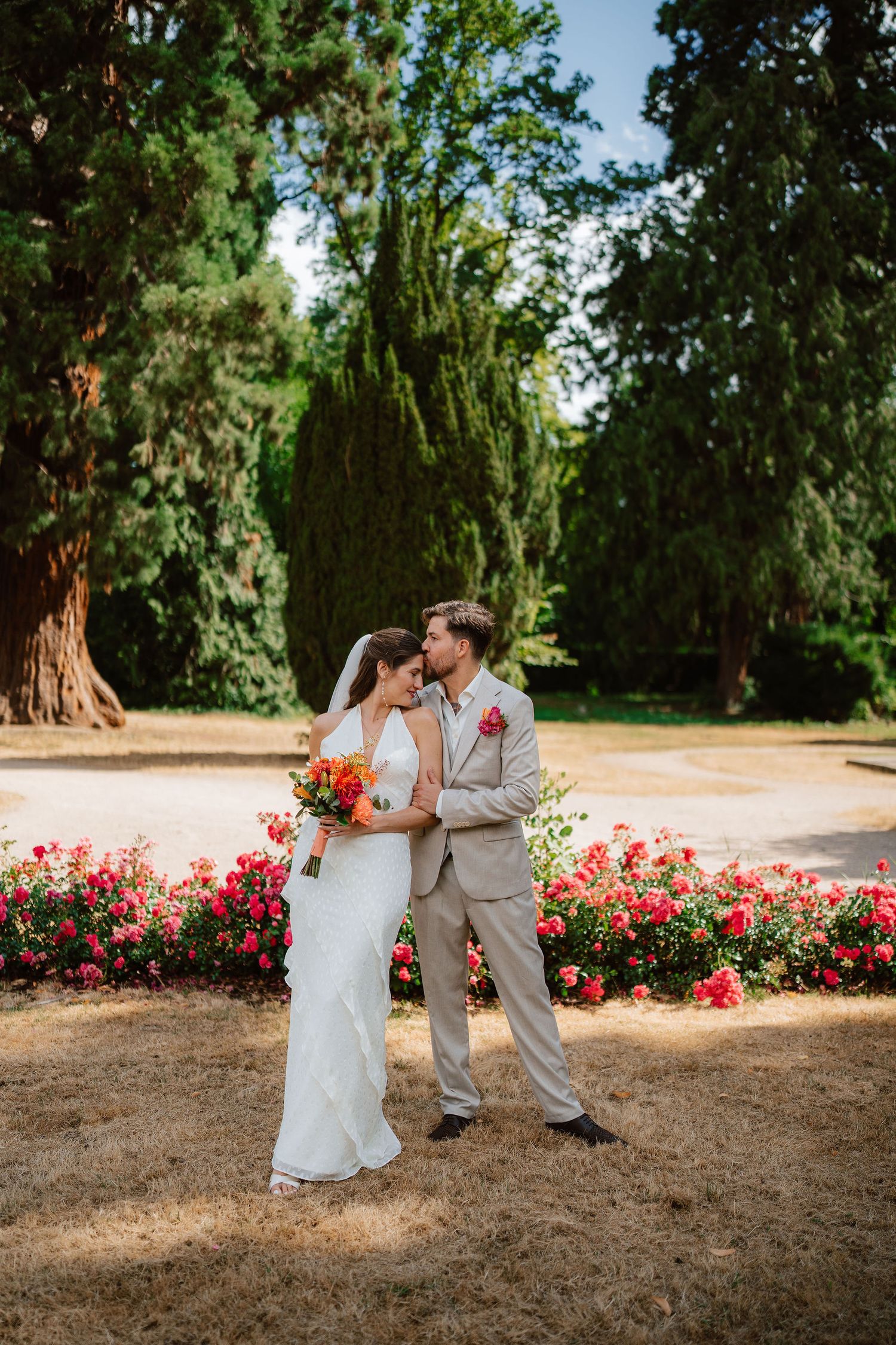 Bride and groom share a romantic kiss in a garden surrounded by vibrant red and orange flowers and tall green trees.