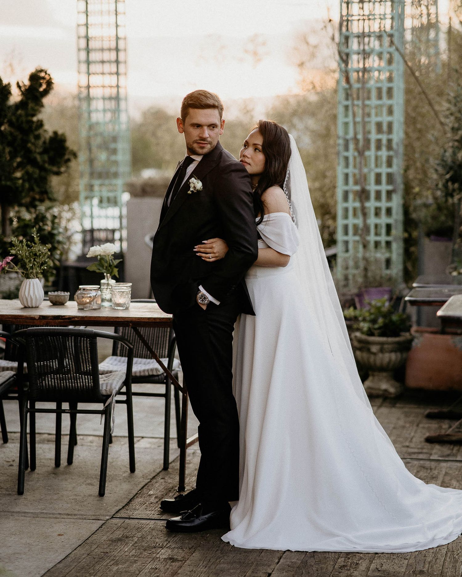A newlywed couple poses together on an outdoor terrace with garden decor and a long flowing white wedding gown.