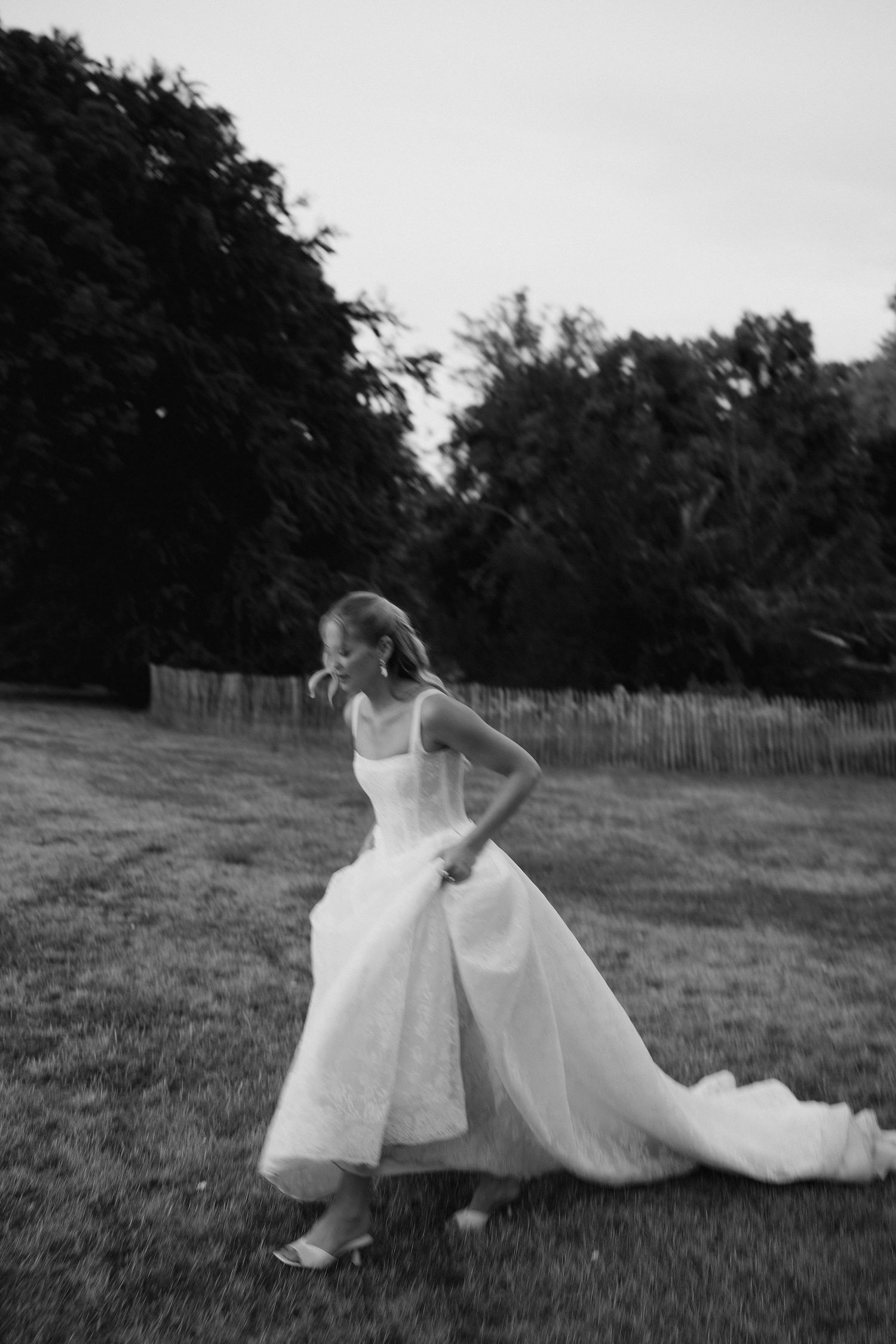 Black and white photo of a bride in a flowing white gown walking gracefully across a lush green lawn.
