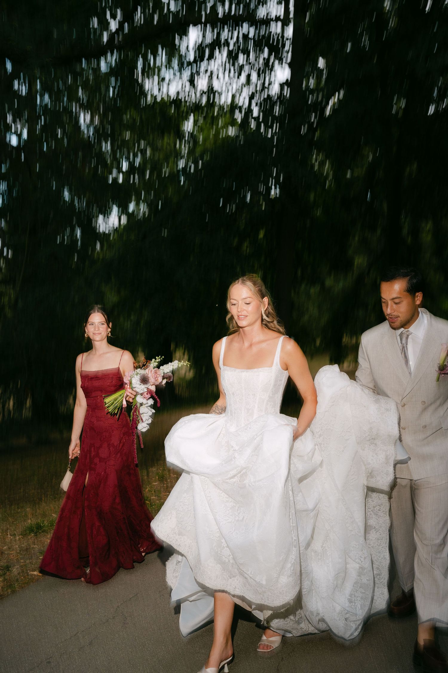 Bride in white gown lifts dress while walking with bridesmaid in burgundy and groomsman outdoors.