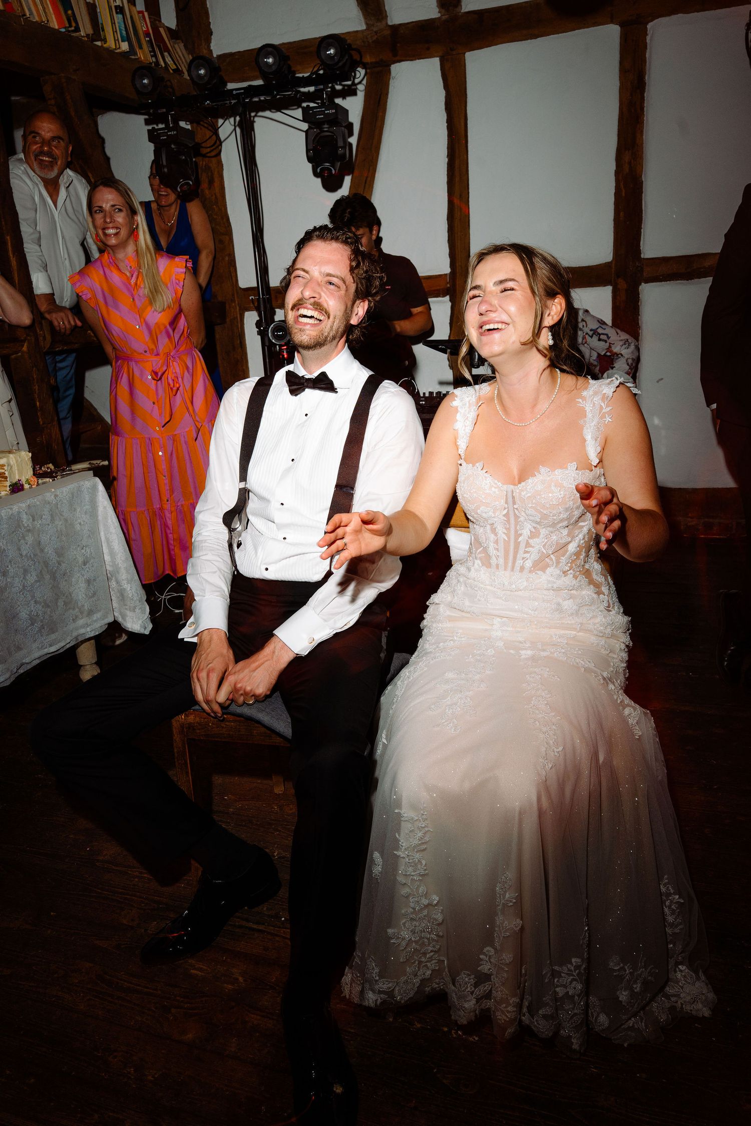 Laughing bride and groom sitting together at wedding reception in rustic barn with guests celebrating behind them.