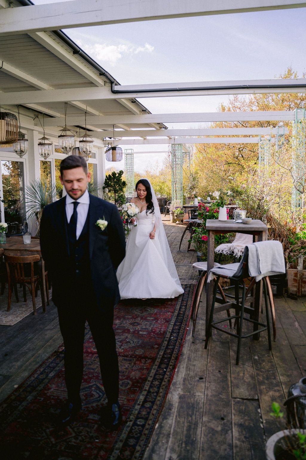 A couple in wedding attire standing on a rustic covered outdoor patio with autumn foliage in the background.
