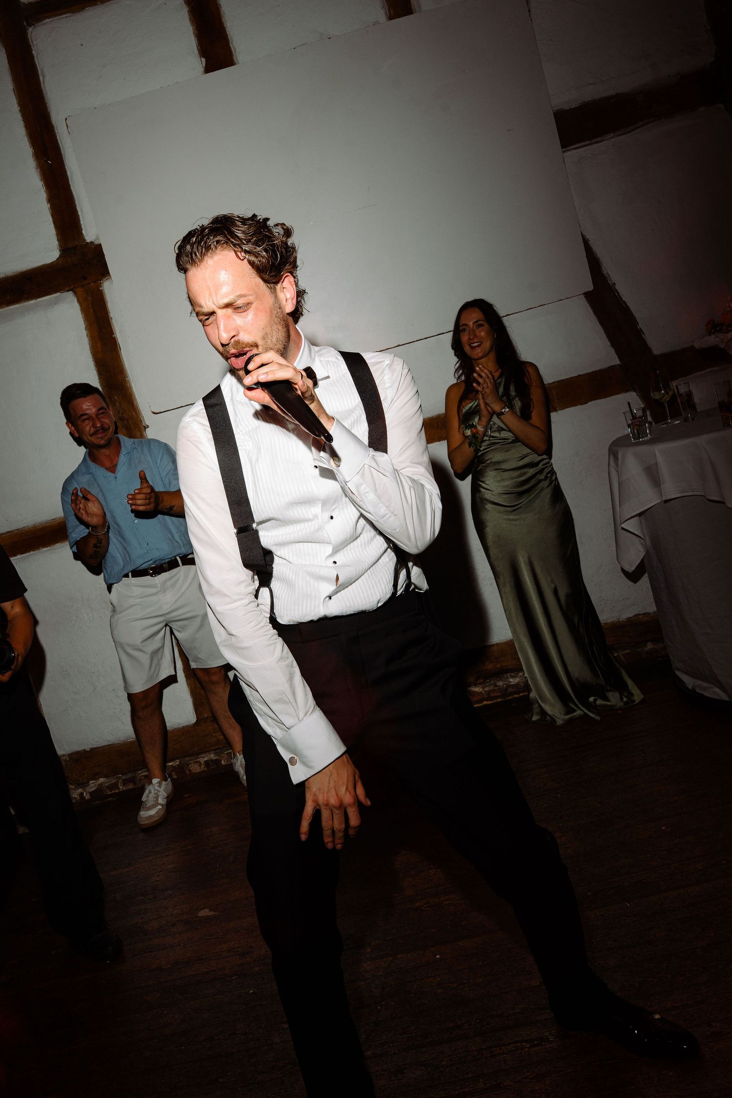 Man in white shirt and suspenders dancing at a wedding reception while guests clap and cheer behind him.
