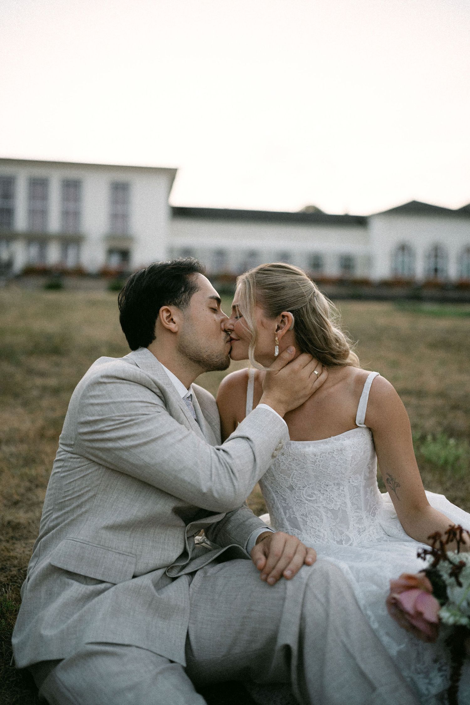 A bride and groom share a romantic kiss while sitting on grass outside a white wedding venue at dusk.