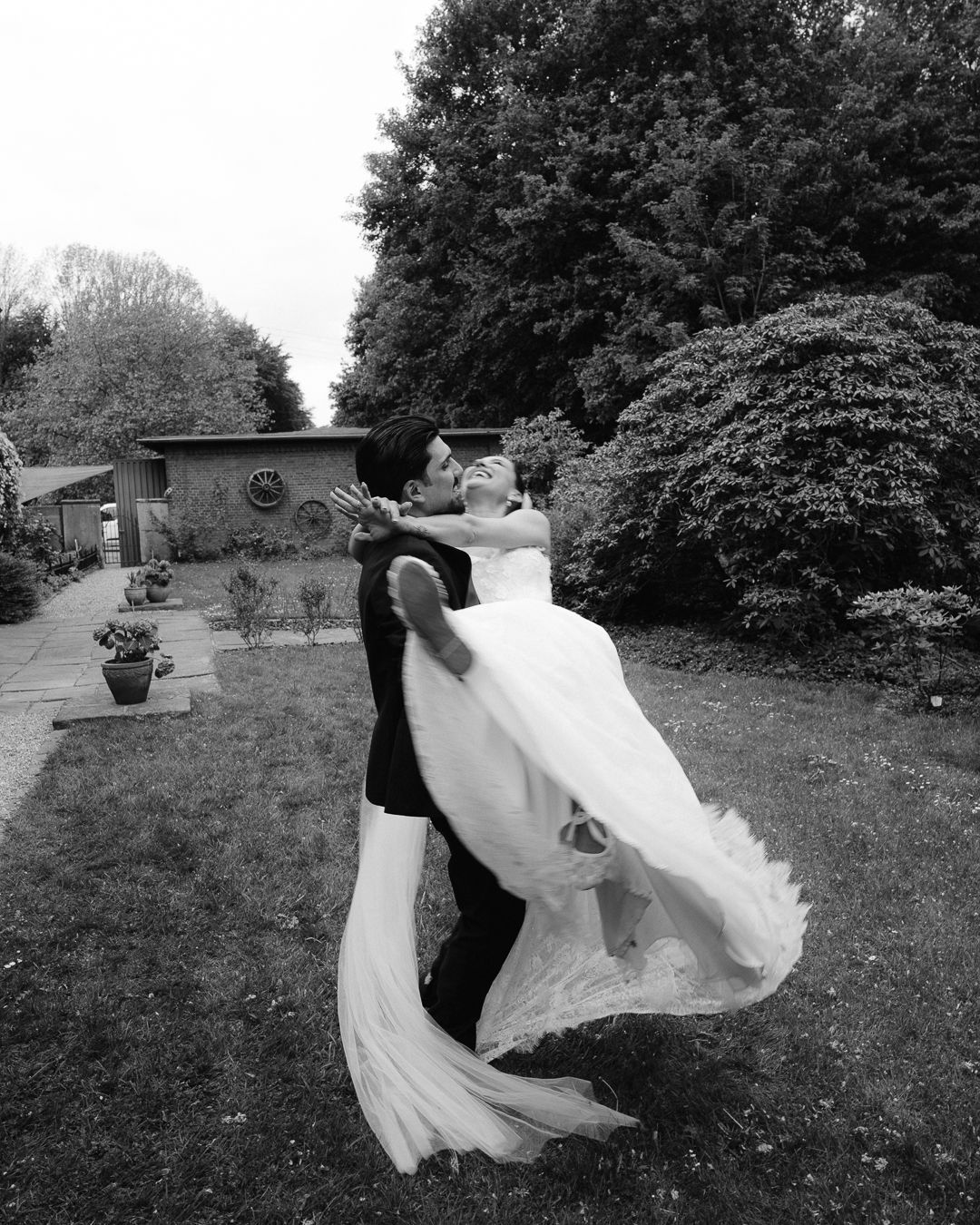 Black and white photo of groom lifting bride in wedding dress outdoors in lush garden setting.