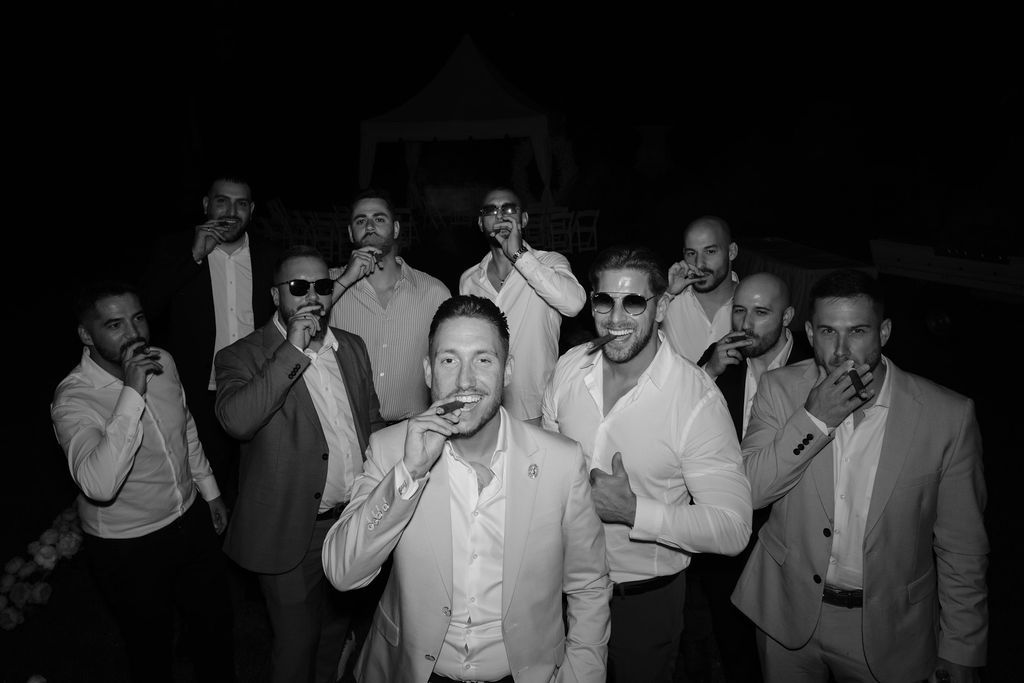 A black and white photo shows a group of men in formal attire smoking cigars together during an evening celebration.