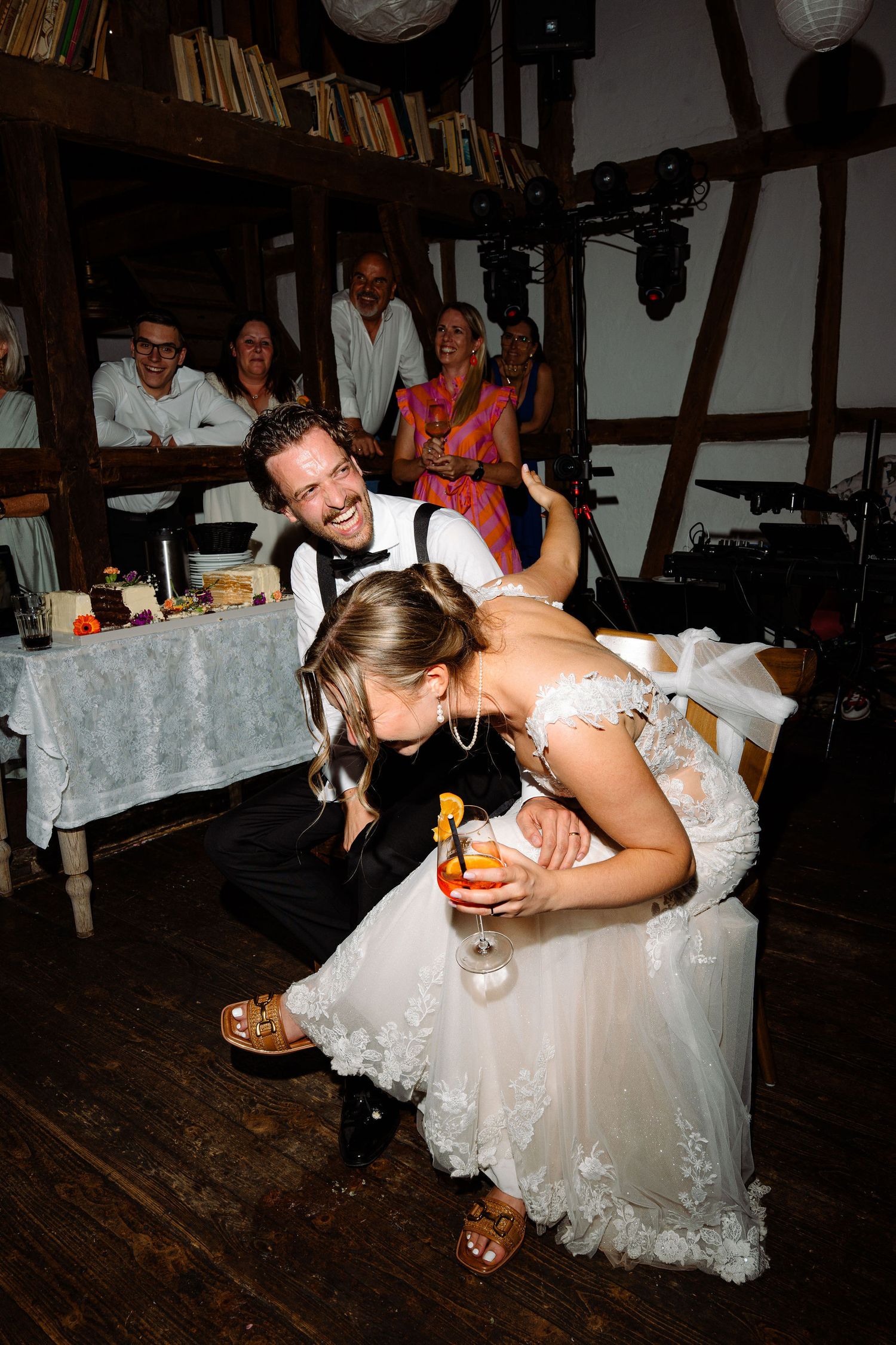 Bride in white lace gown and groom in black suit laugh together at wedding reception with guests in background.