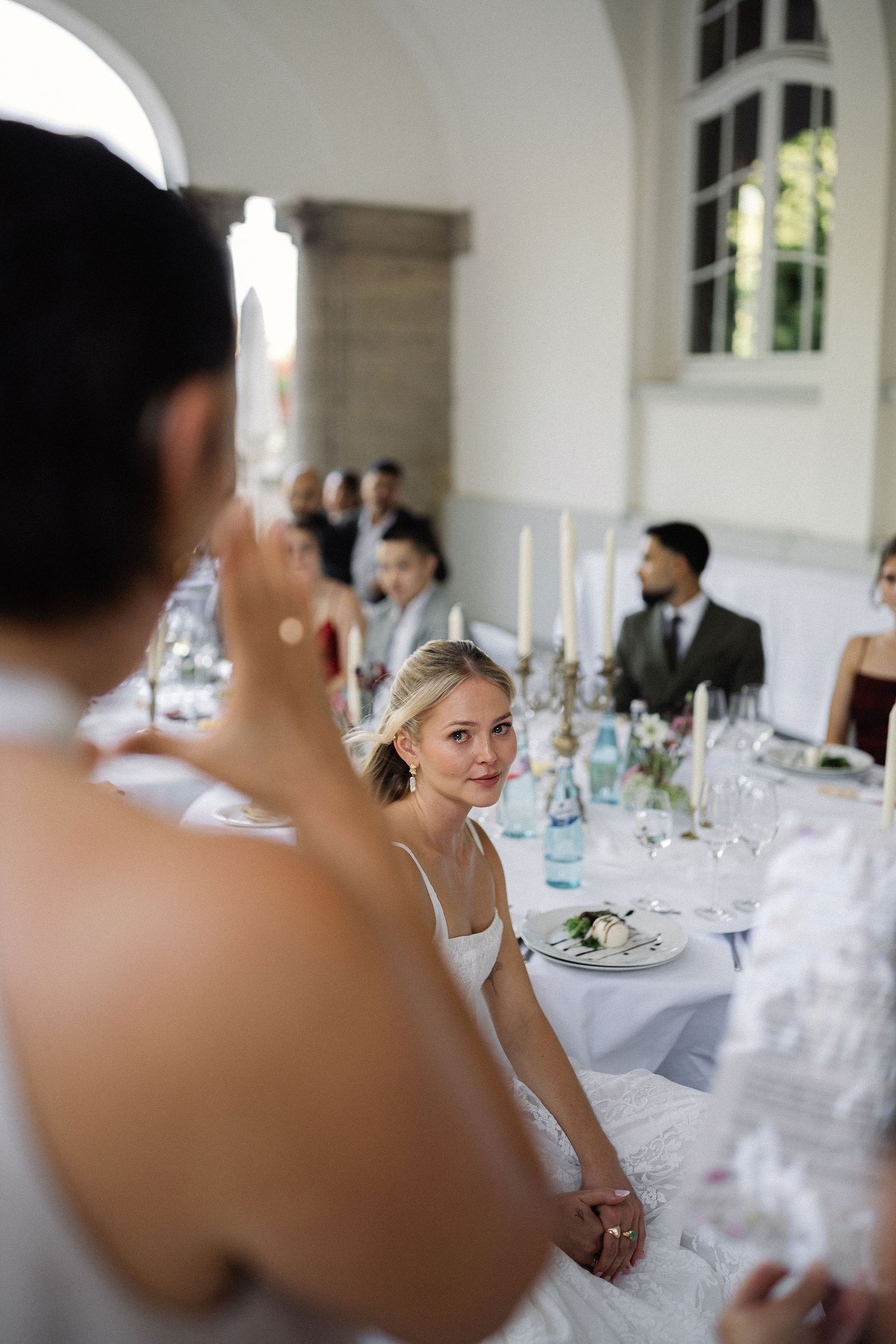 Bride in white dress sits at elegant wedding reception table with candles, flowers, and guests in formal attire.