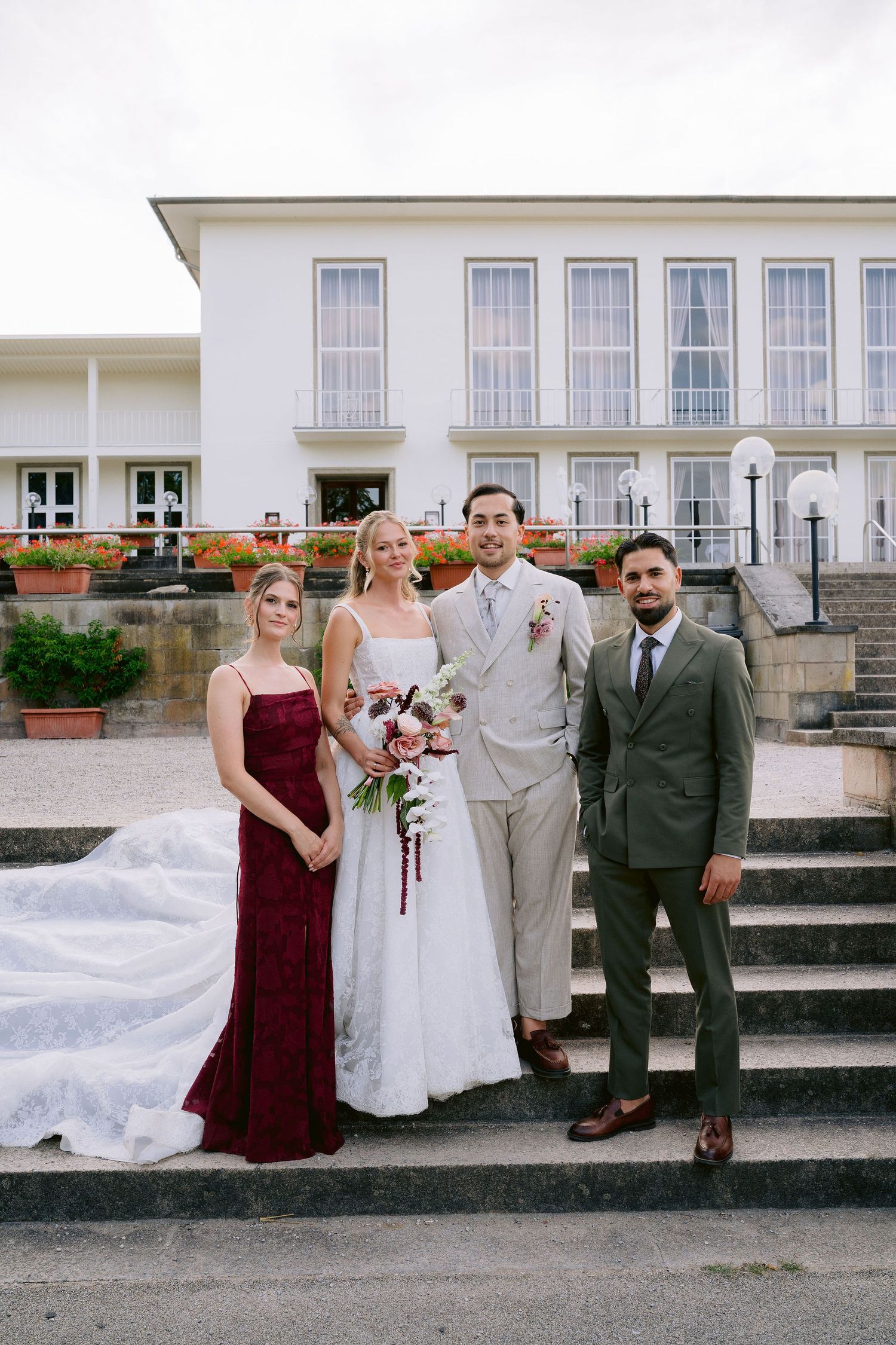 Wedding party of four poses on steps outside white venue, bride in white gown holding bouquet with bridesmaid in burgundy dress.