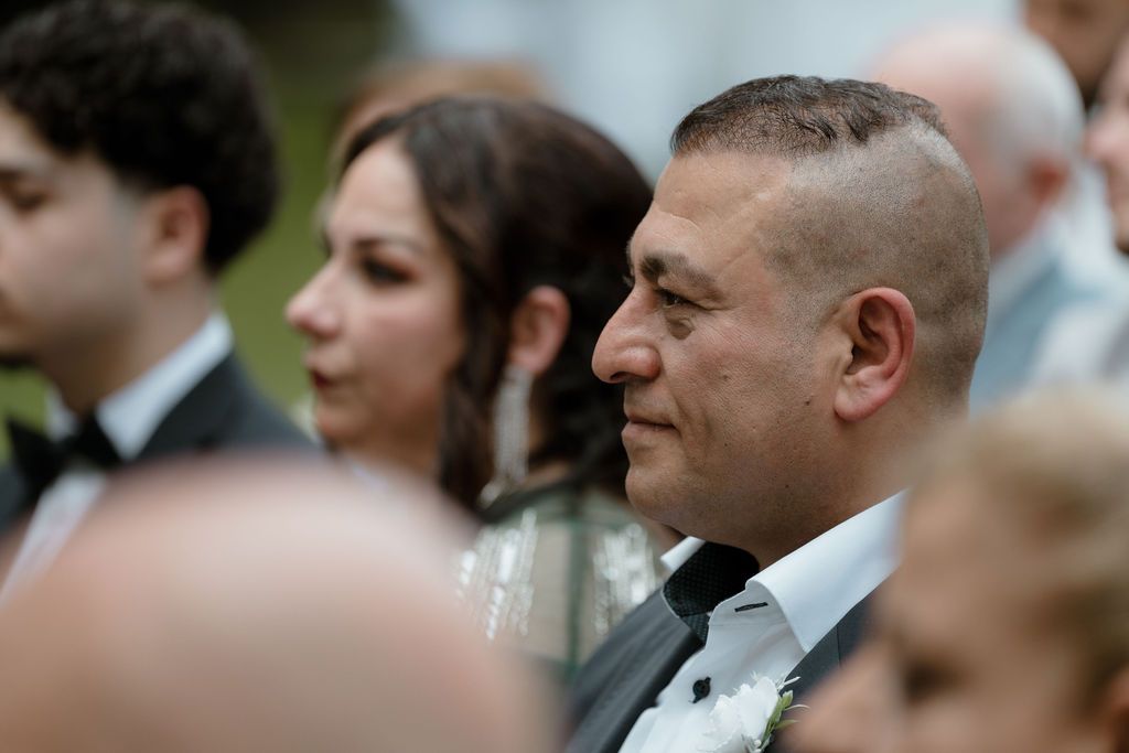 Guests are seated outdoors in formal attire at a wedding ceremony on a sunny day.