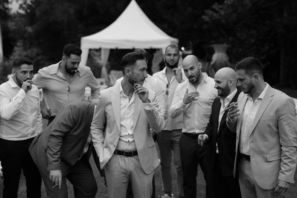 Black and white photo of groomsmen sharing a candid moment outdoors under a white tent.