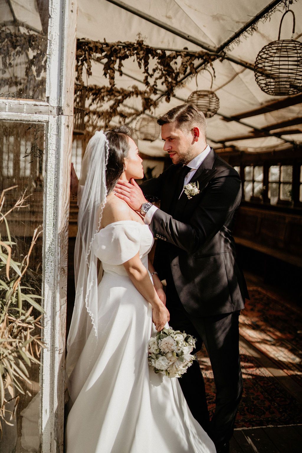 A bride and groom sharing an intimate first look moment in a rustic greenhouse setting with natural light streaming through.
